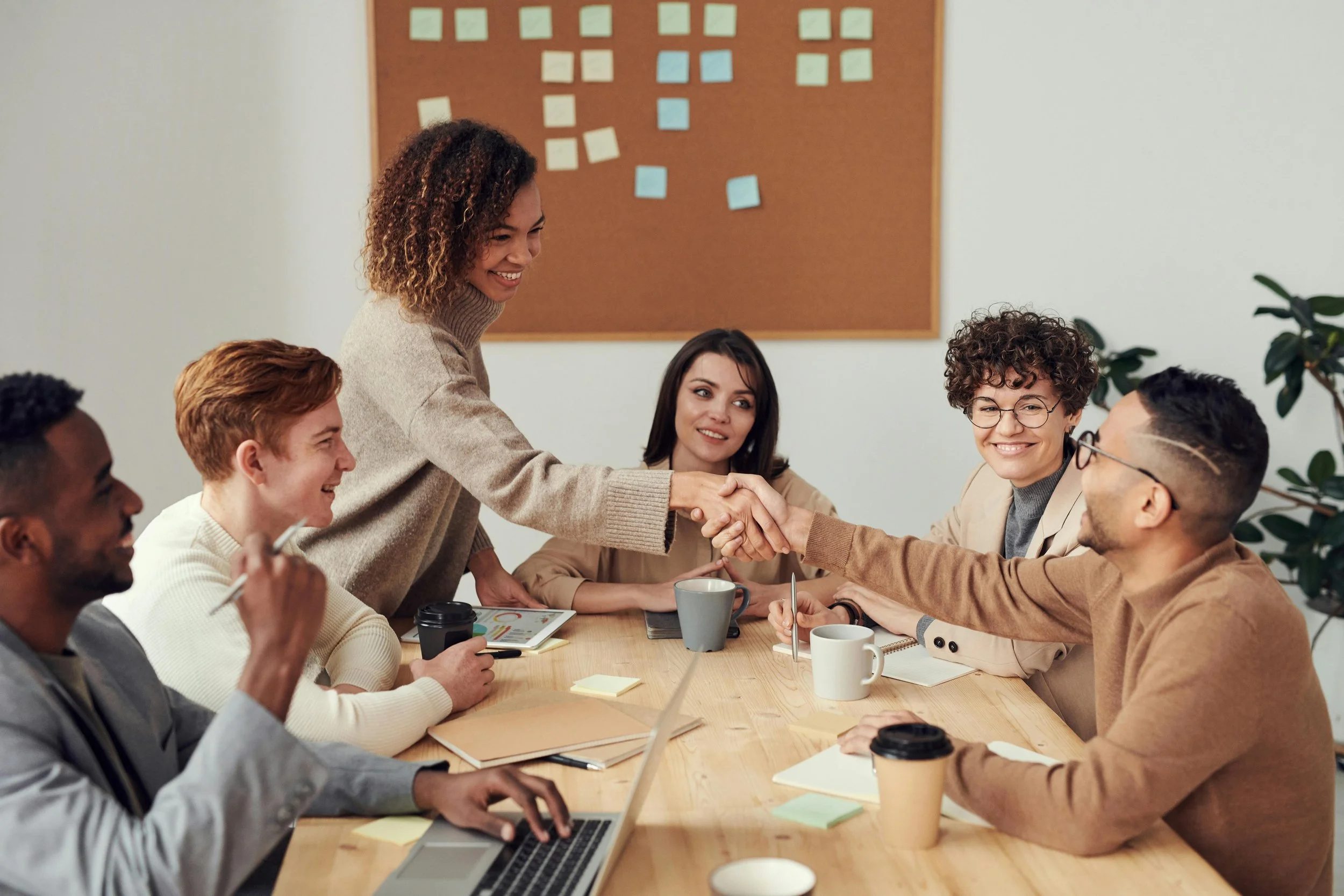 Group of diverse professionals in a meeting room, smiling and shaking hands over a wooden conference table with laptops, notebooks, coffee cups, and colorful sticky notes.