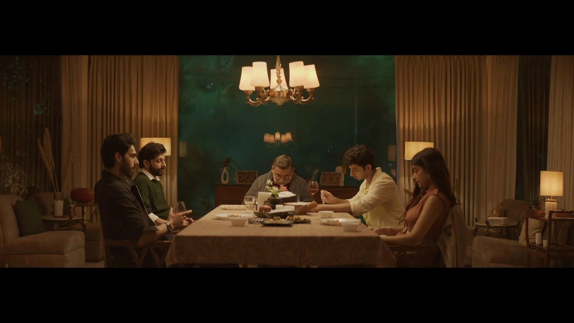 Six people seated around a dining table in a warmly lit room with golden curtains and lamps, engaged in a meal and conversation.