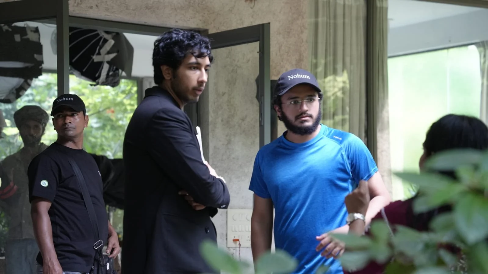 Four men engaged in conversation indoors. Two men in the foreground are listening attentively—one in a black suit with crossed arms, the other in a blue t-shirt and cap labeled 'Nohum'. Two men are in the background, one in a black shirt and cap labeled 'Nohum', and another partially visible.