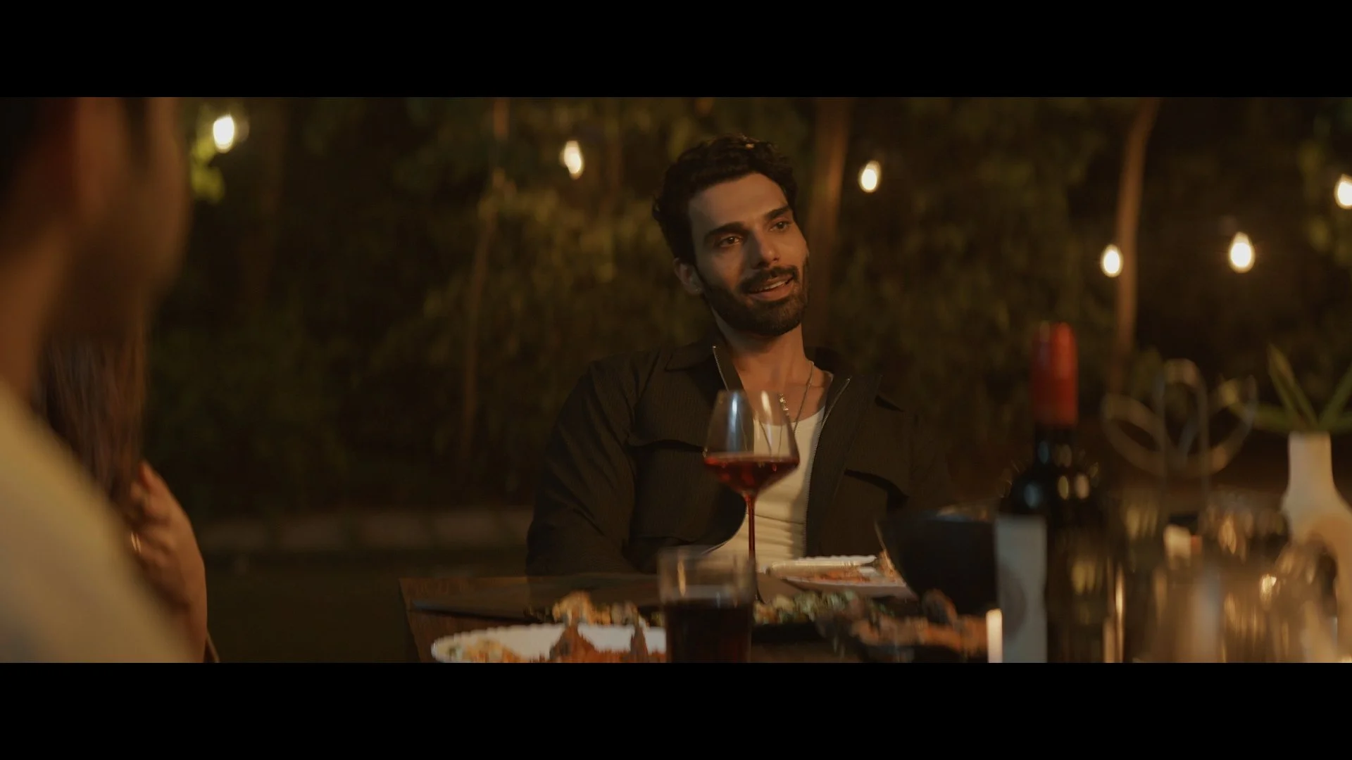 Group of people dining outdoors at night with string lights overhead, a man is smiling and talking, drinks and food on the table.