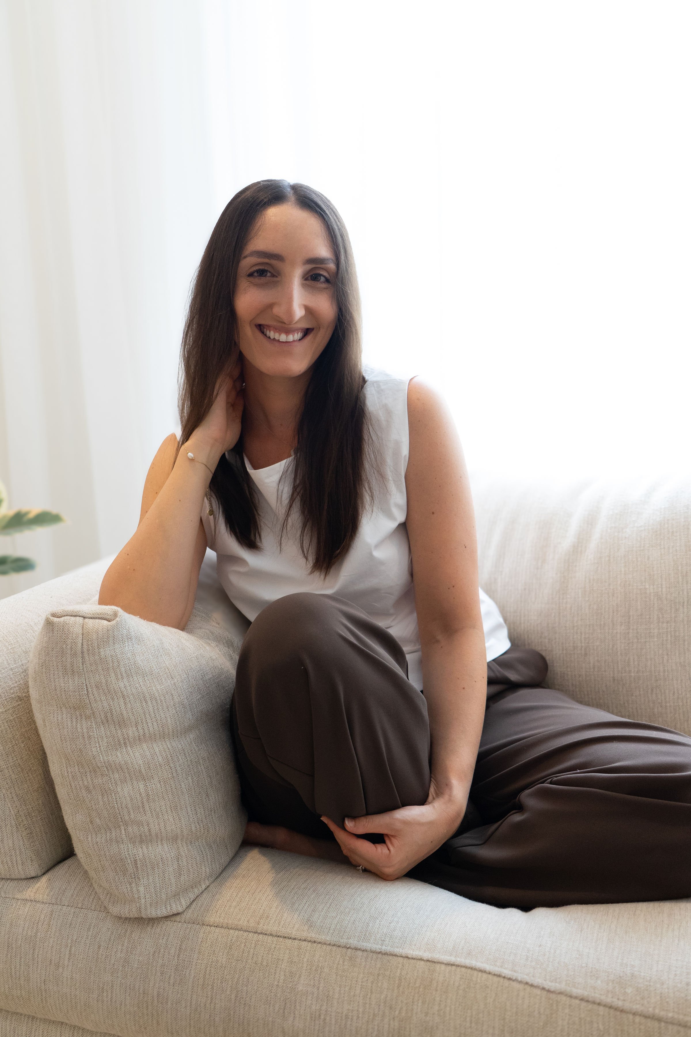 A woman with long dark hair, smiling, sitting on a beige sofa in a bright room with white curtains.