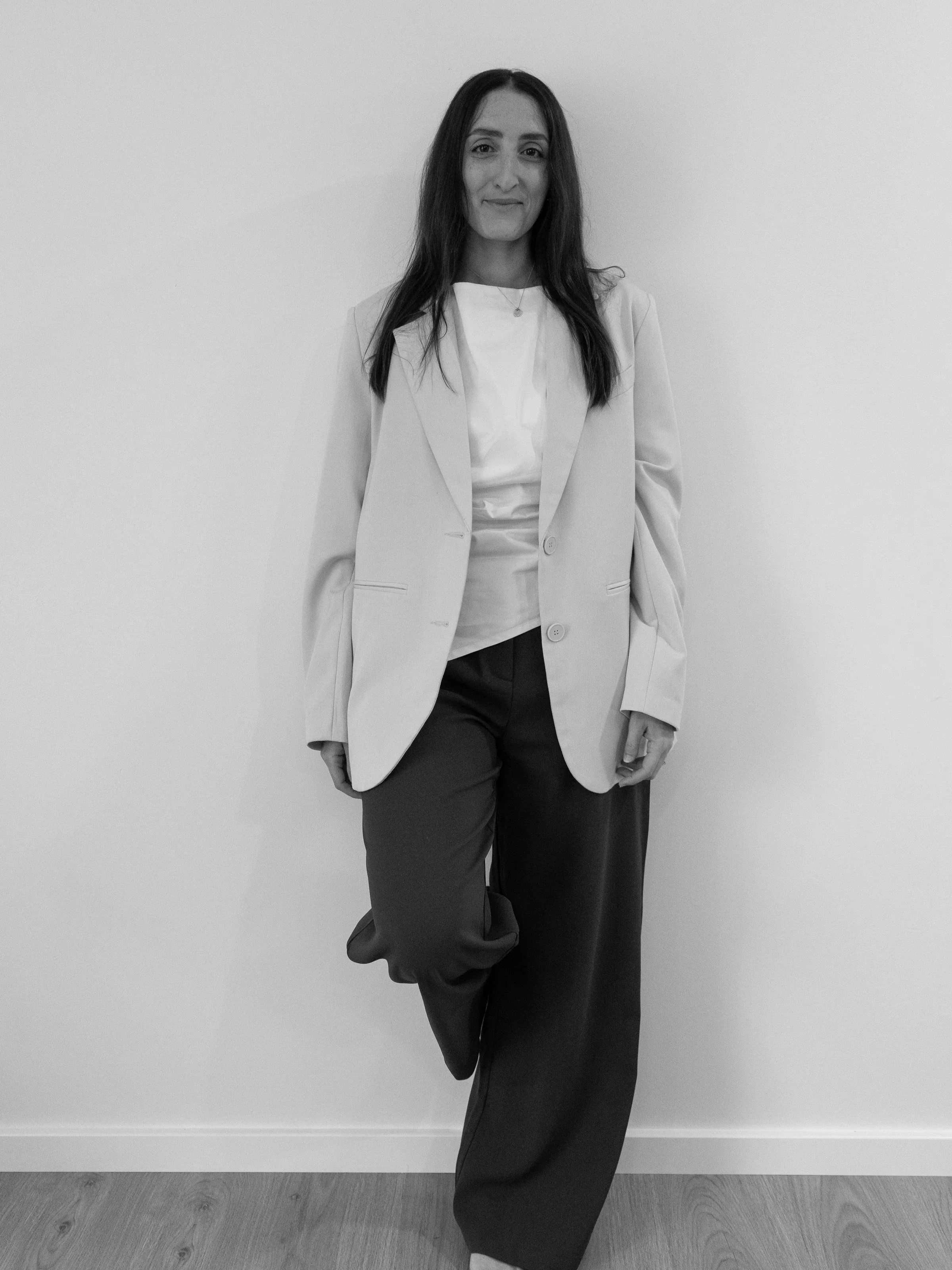 A woman with long dark hair standing against a plain white wall, wearing a light-colored blazer, a white top, and dark wide-legged pants.