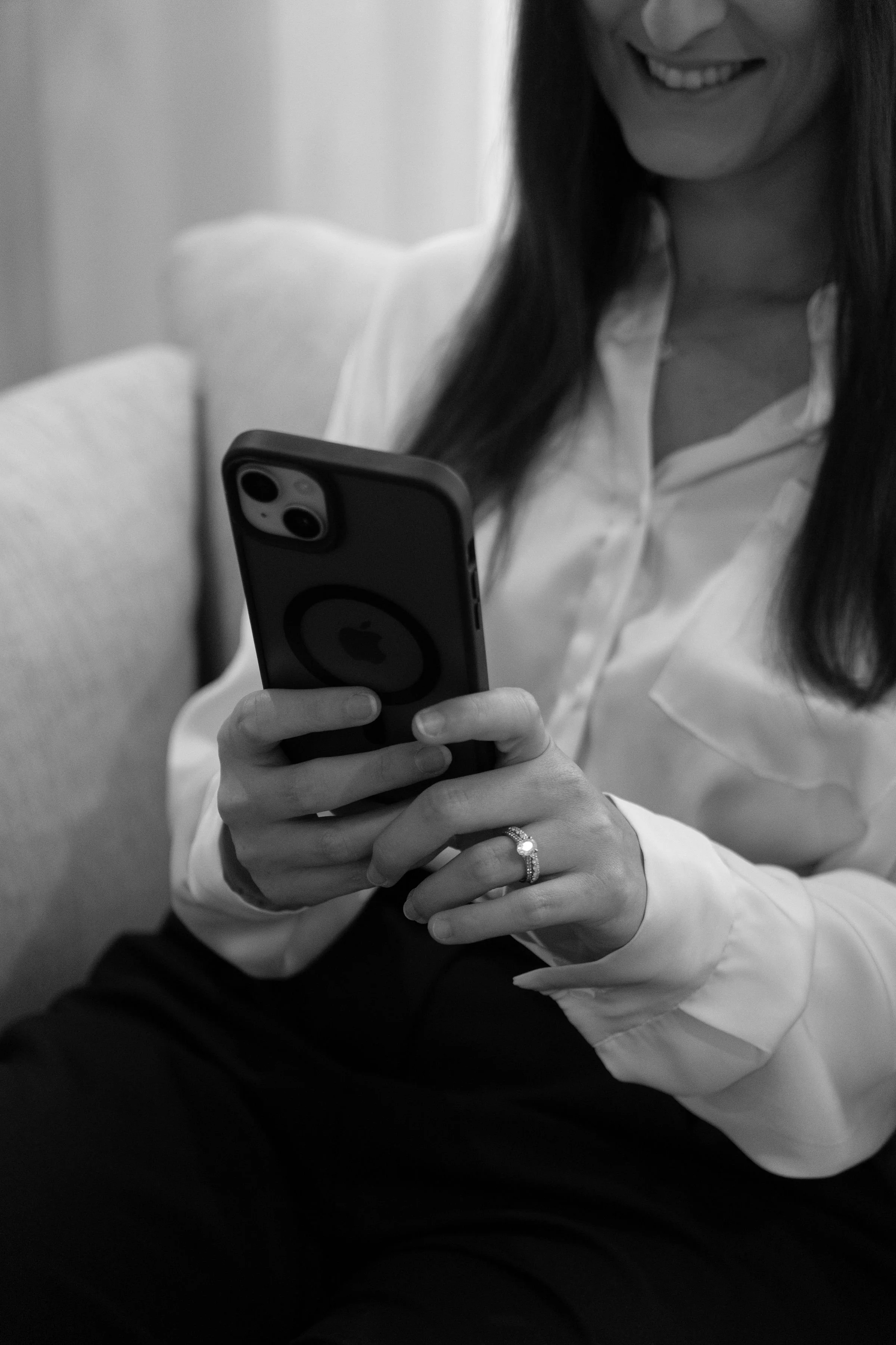 A woman with long hair, smiling, wearing a white blouse, sitting on a light-colored sofa, holding a smartphone with a circular grip, wearing a ring on her finger.