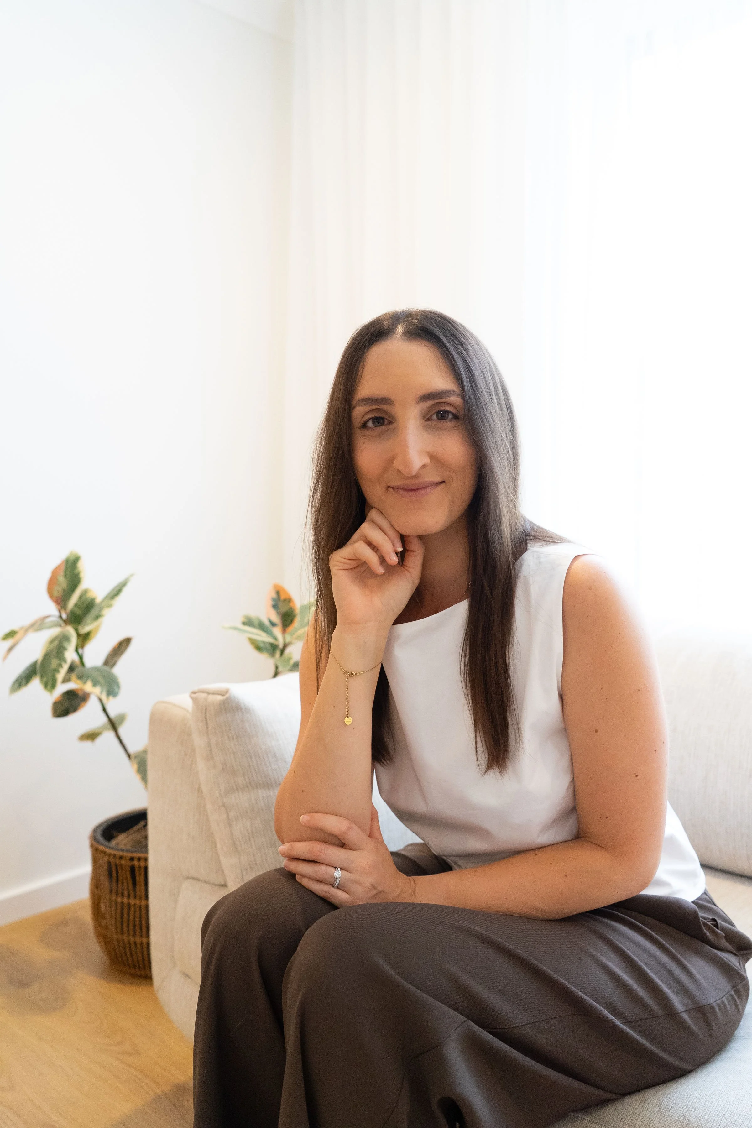 A woman sitting on a beige sofa, smiling, with a hand resting on her chin, in a bright room with a potted plant in the background.