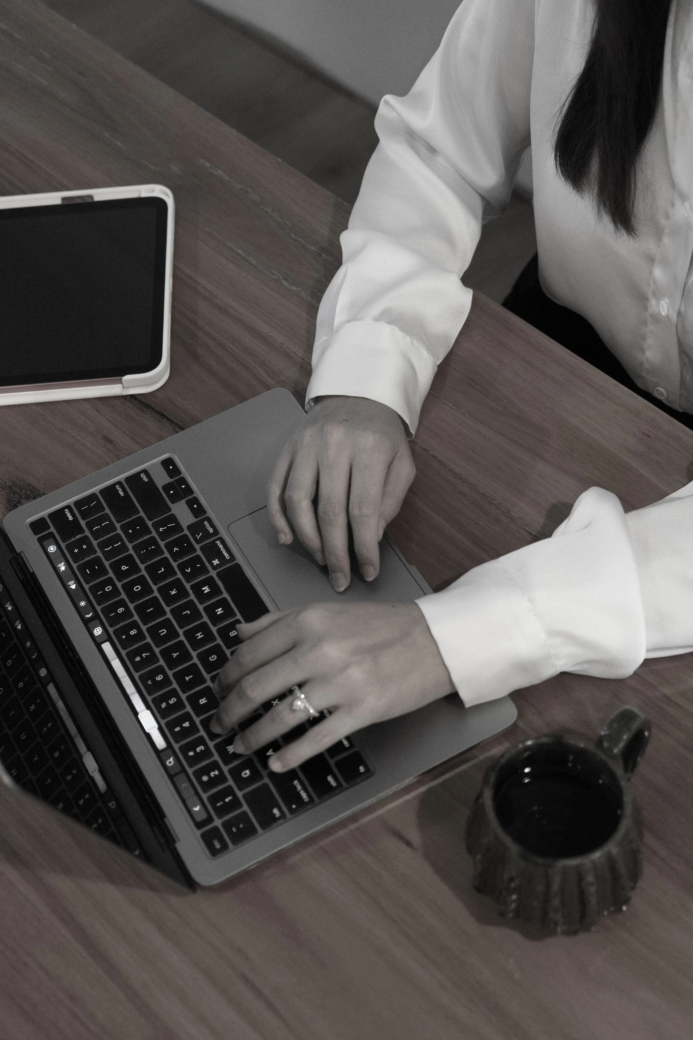 A person wearing a white shirt working on a silver laptop at a wooden desk, with a cup of black coffee and a tablet nearby.