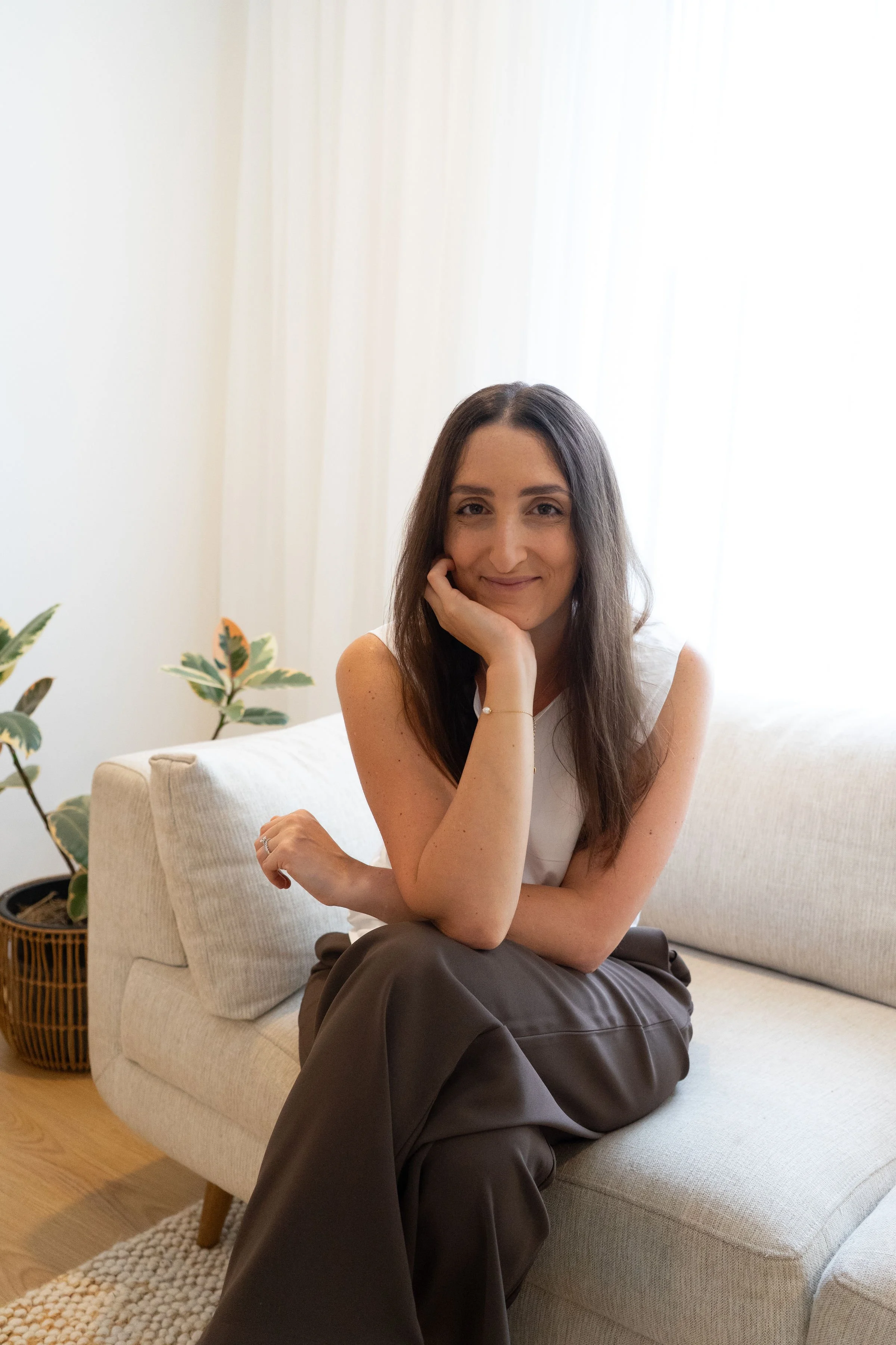 A woman sitting on a light-colored sofa in a bright room, smiling and resting her chin on her hand, with green plants in the background.