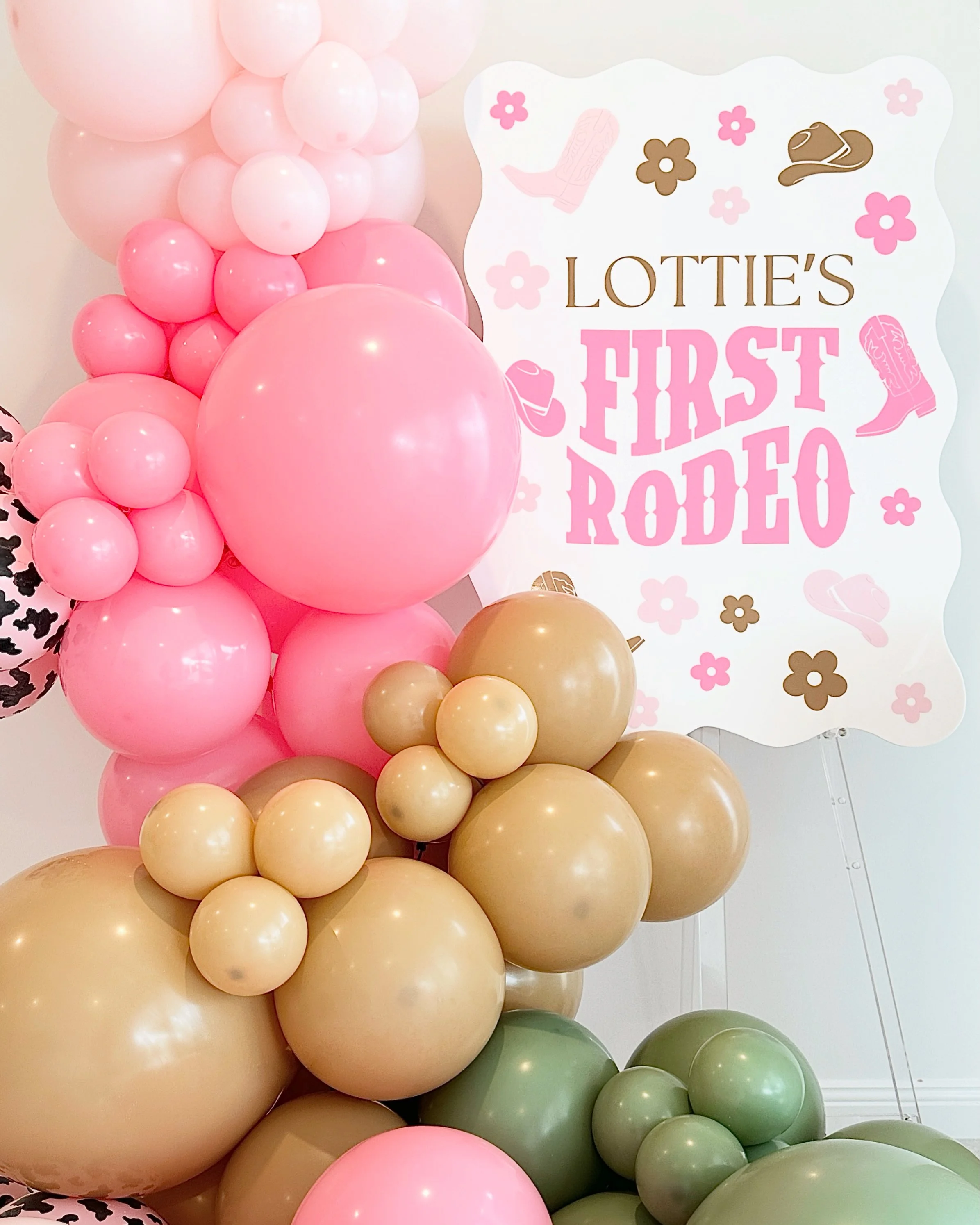 A large arrangement of pink, beige, and green balloons near a sign that reads 'Lottie's First Rodeo' with illustrations of cowboy boots, hats, and flowers.