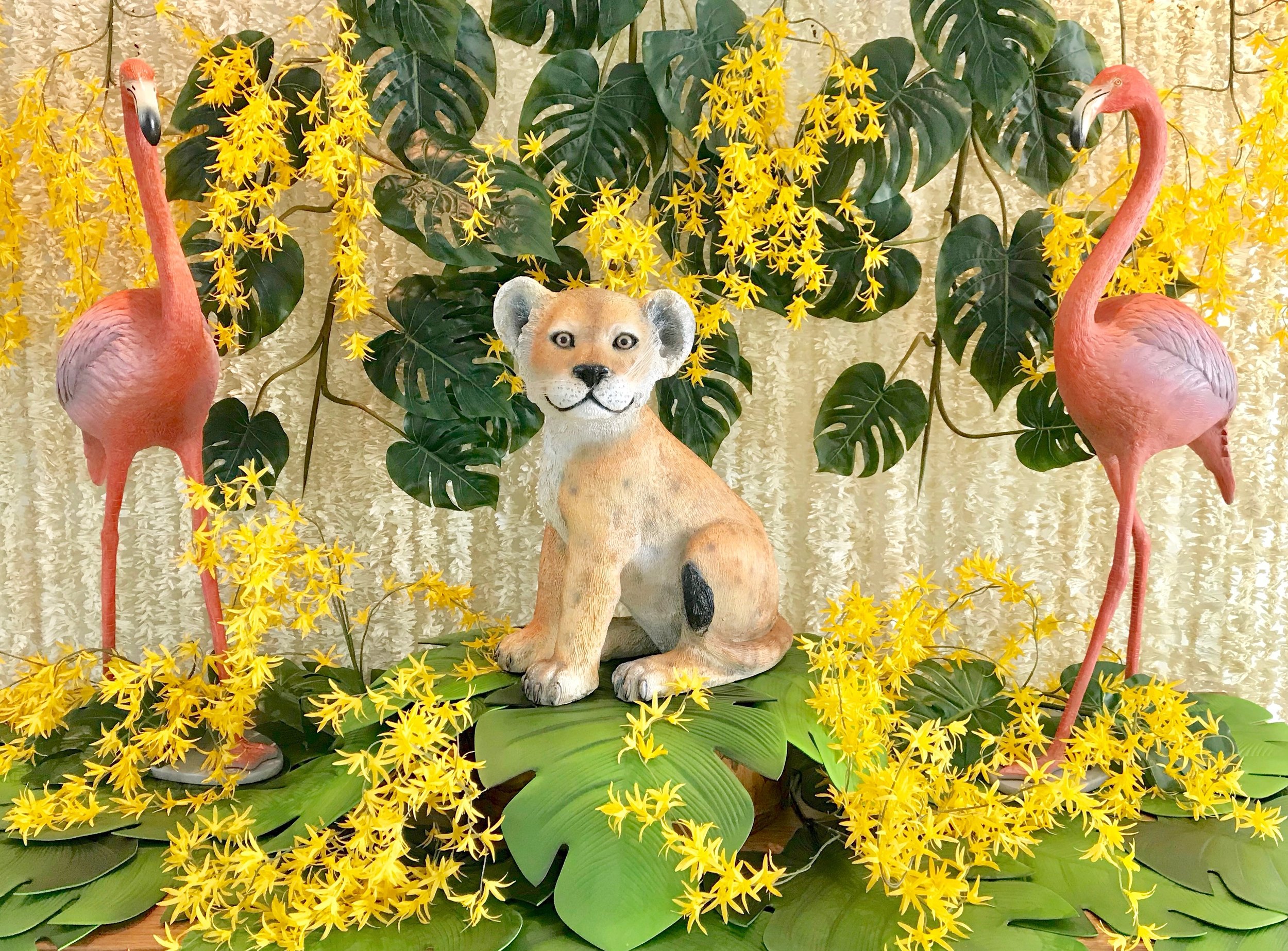 Decorative display featuring two pink flamingo statues and a lion cub figurine, surrounded by green leaves and yellow flowers.