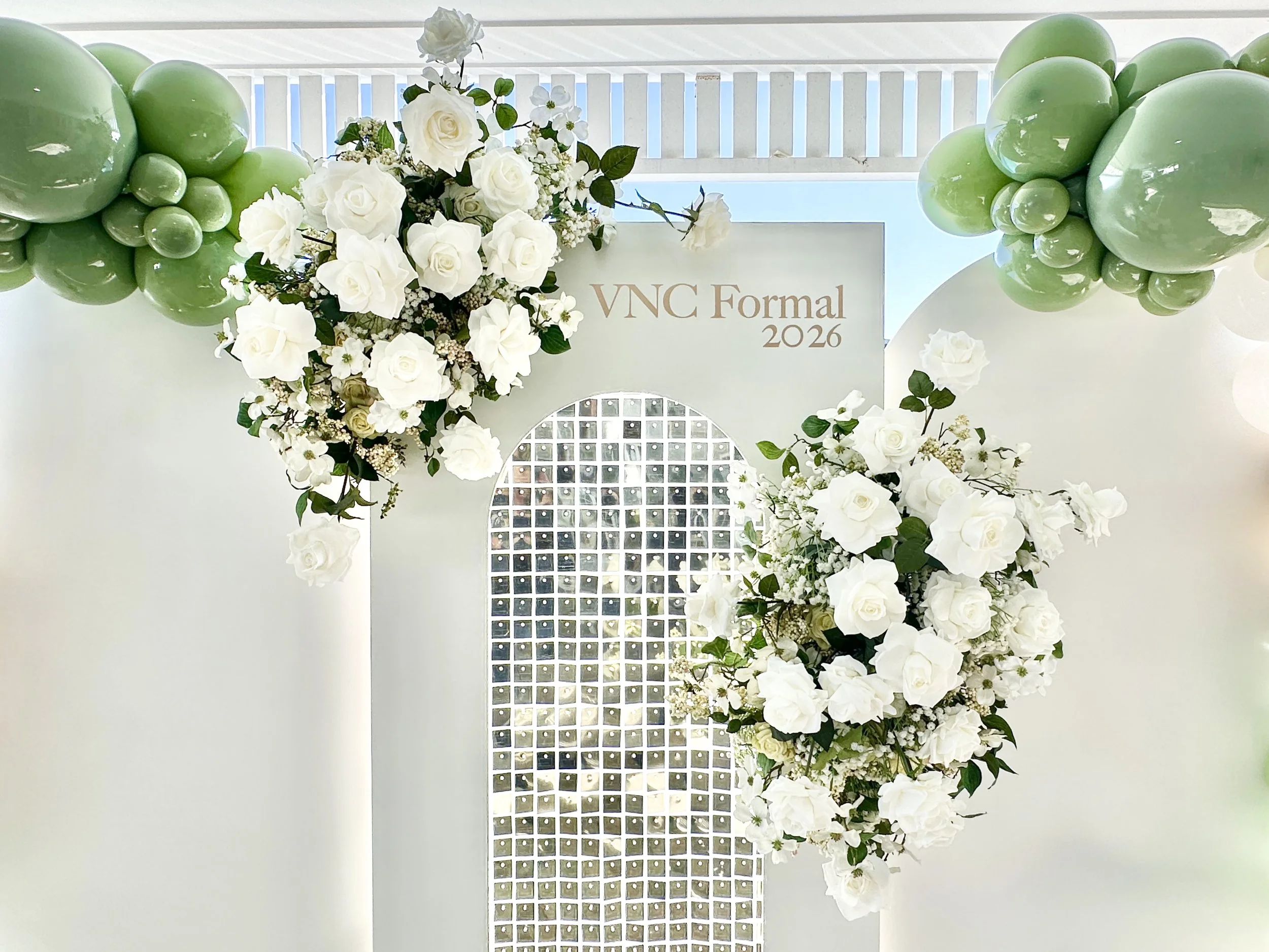Decorative white flower arrangement with roses and greenery on a backdrop of green balloons and a white lattice structure, with a sign reading 'VNC Formal 2026' in the background.
