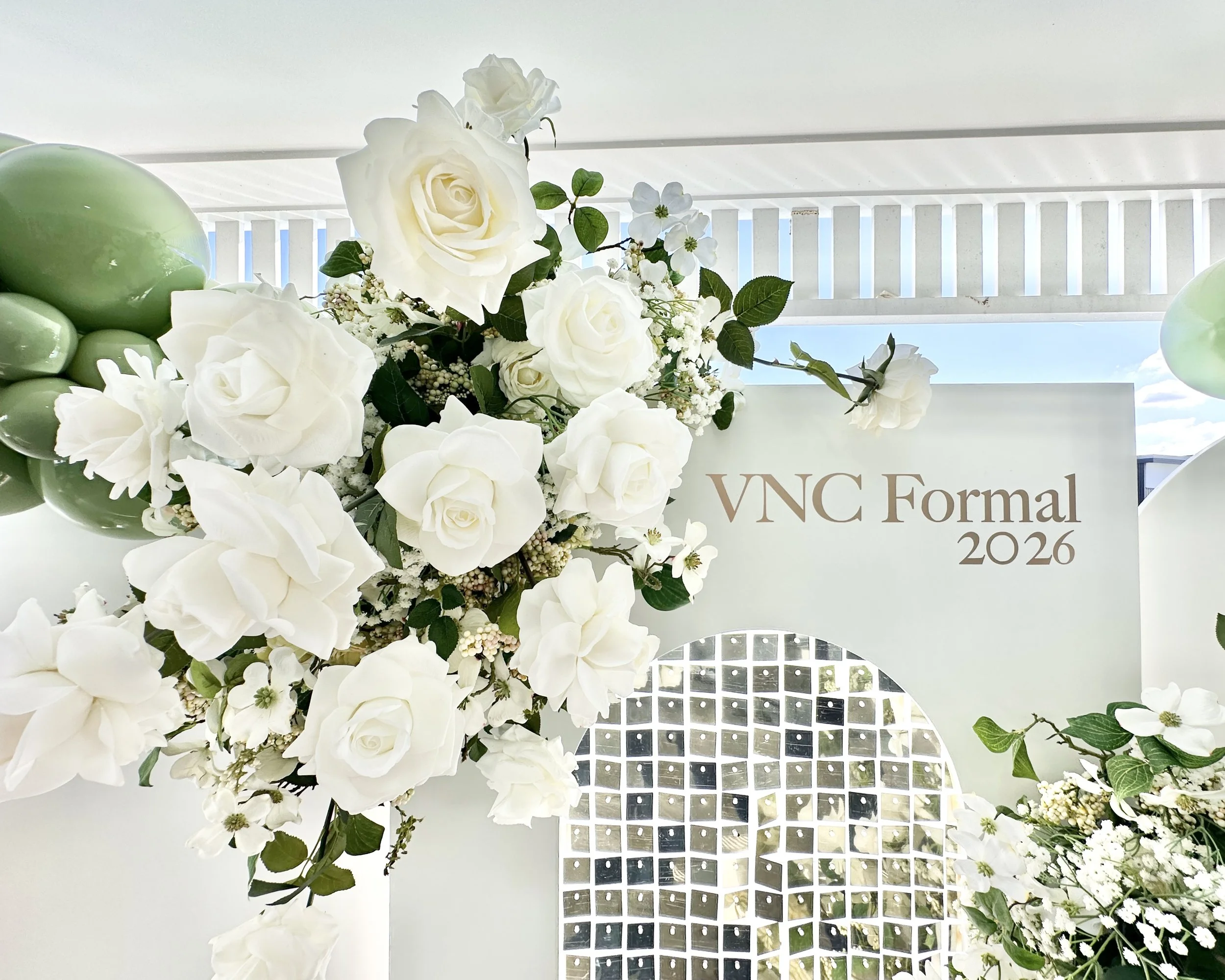A floral arrangement with white roses, small white flowers, and green leaves, decorated with green balloons, set against a white backdrop with a blue sky visible through a window, and a sign that reads "VNC Formal 2026".