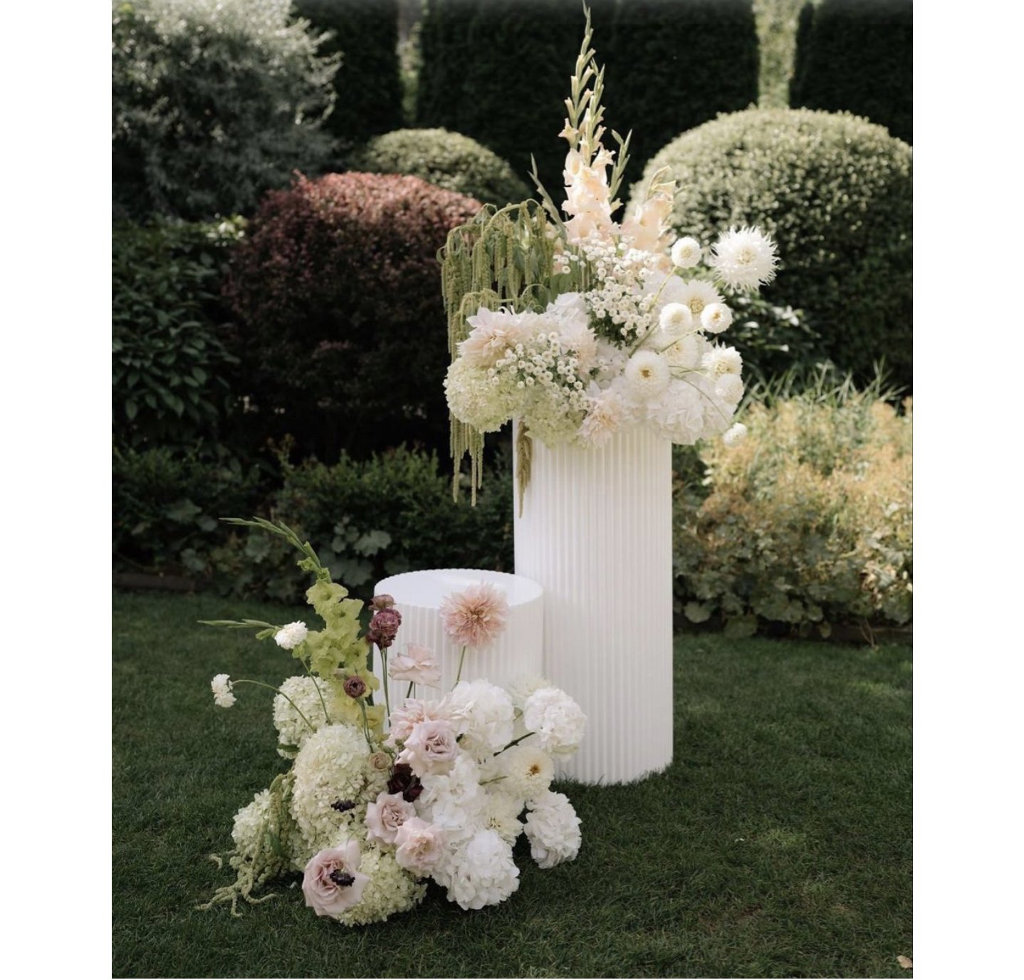 Elegant outdoor floral arrangement with white and soft pink flowers on and around white pedestal stands, set in a garden with lush greenery and shrubs.
