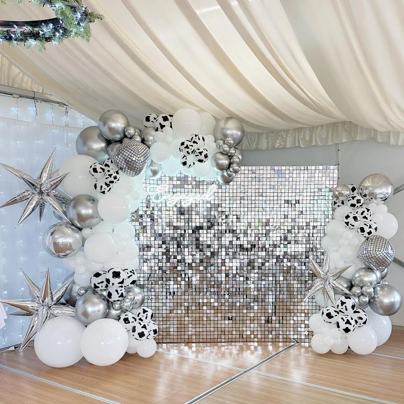 Balloon arch with black and white cow pattern, metallic silver, and white balloons with star-shaped decorations and a sequin backdrop.