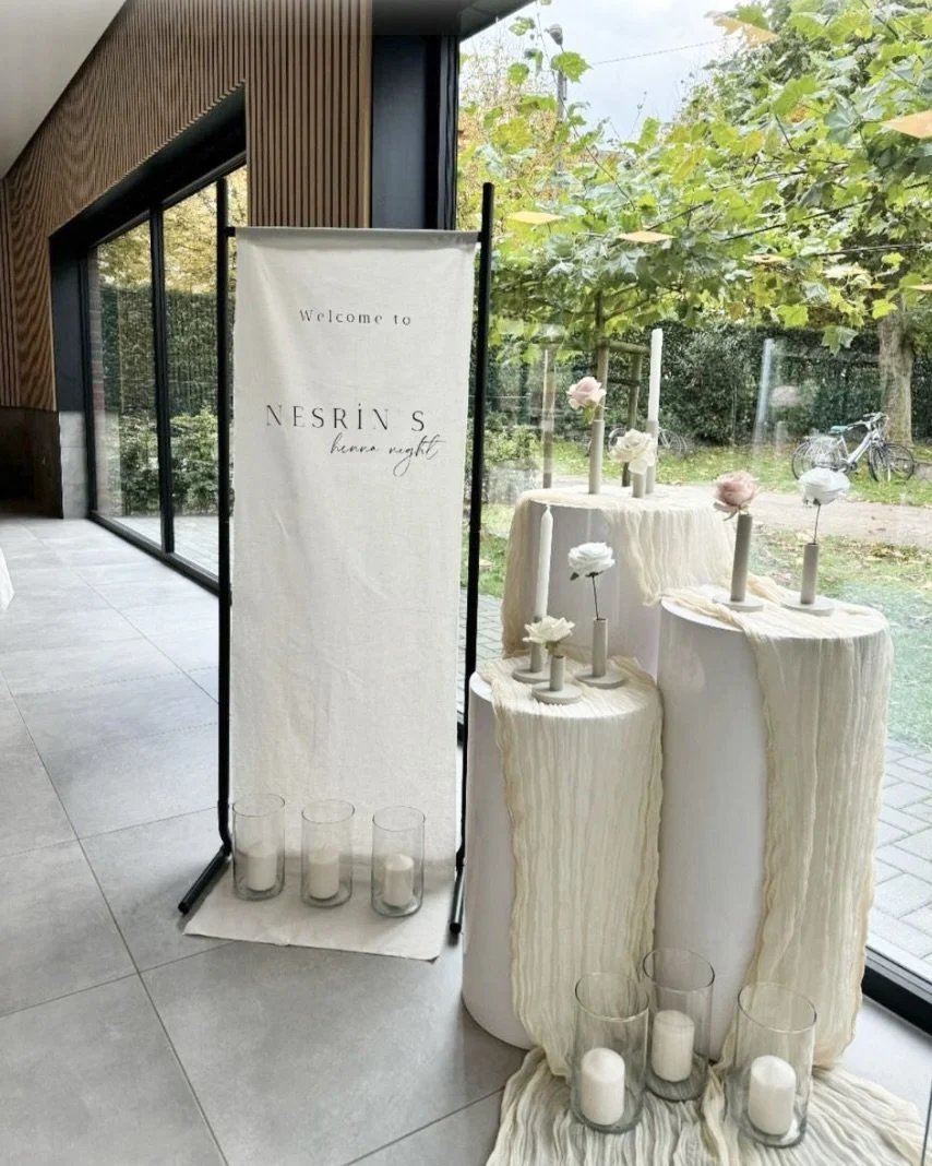 Event display with white candles in glass holders, flowers on candles, and a sign with the text 'Welcome to NESRIV'S bonnie night' near a glass wall overlooking a garden with trees and a bicycle.