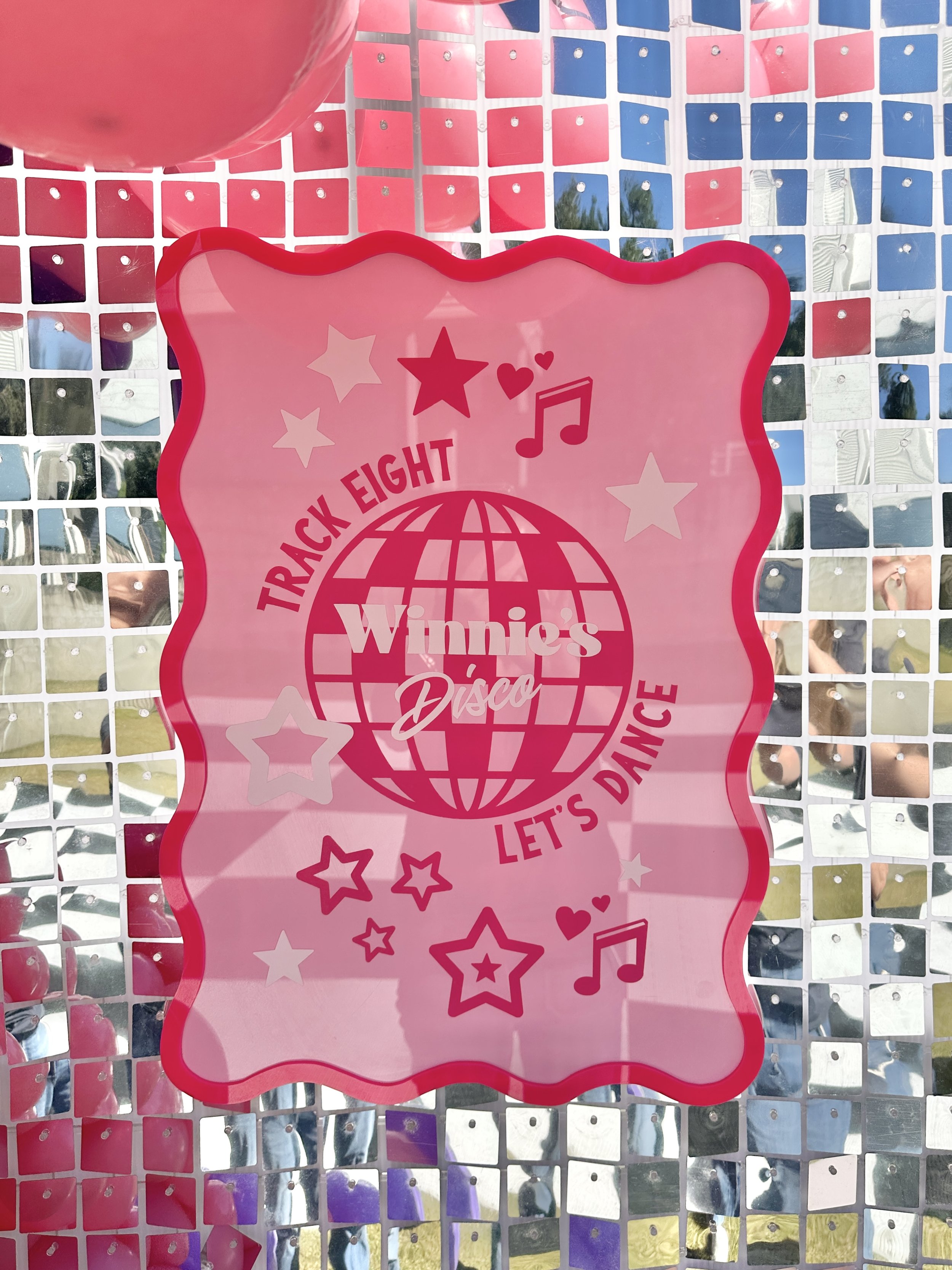 Pink sign with stars, musical notes, a globe, and the text 'Winnie's Disco' and 'Let's dance' on a reflective, grid-like surface. Part of a pink balloon is visible at the top left.