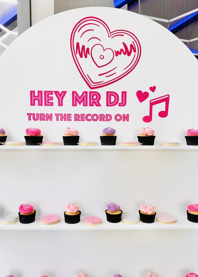 Display of cupcakes with pink and purple frosting on white shelves beneath a sign reading 'Hey Mr DJ, Turn the Record On' with a heart and music note graphic.