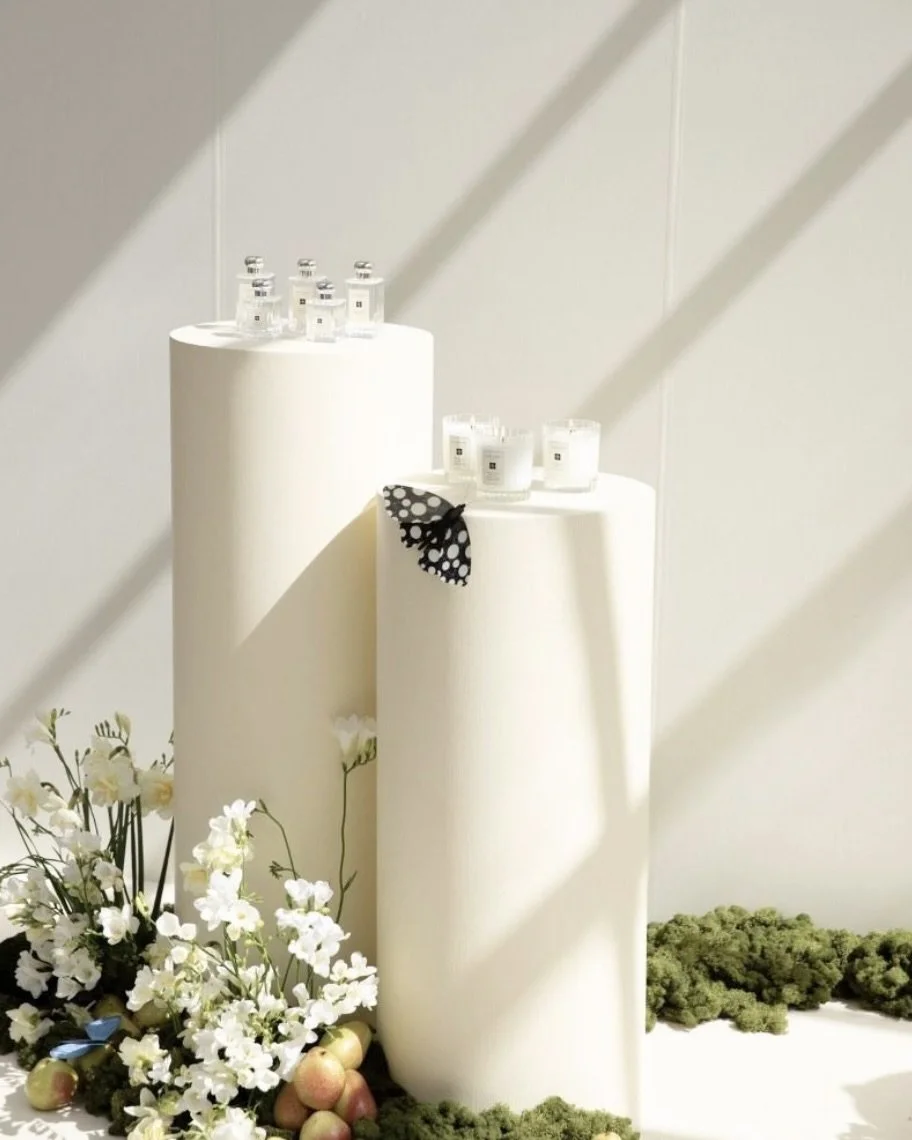 White cylindrical display stands with small bottles and candles, decorated with flowers and apples, and a butterfly-shaped accessory, set against a white background with shadows.