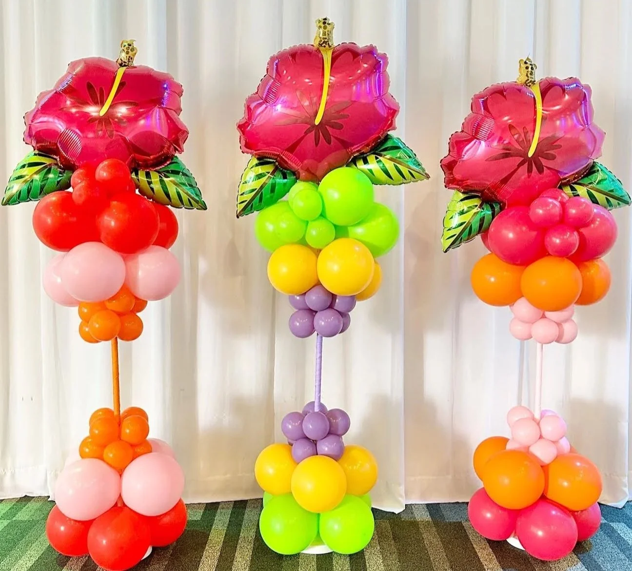 Colorful balloon floral arrangements with large foil balloon flowers on top, featuring pink petals and yellow centers, arranged on stands with clusters of smaller balloons in red, pink, orange, purple, and yellow