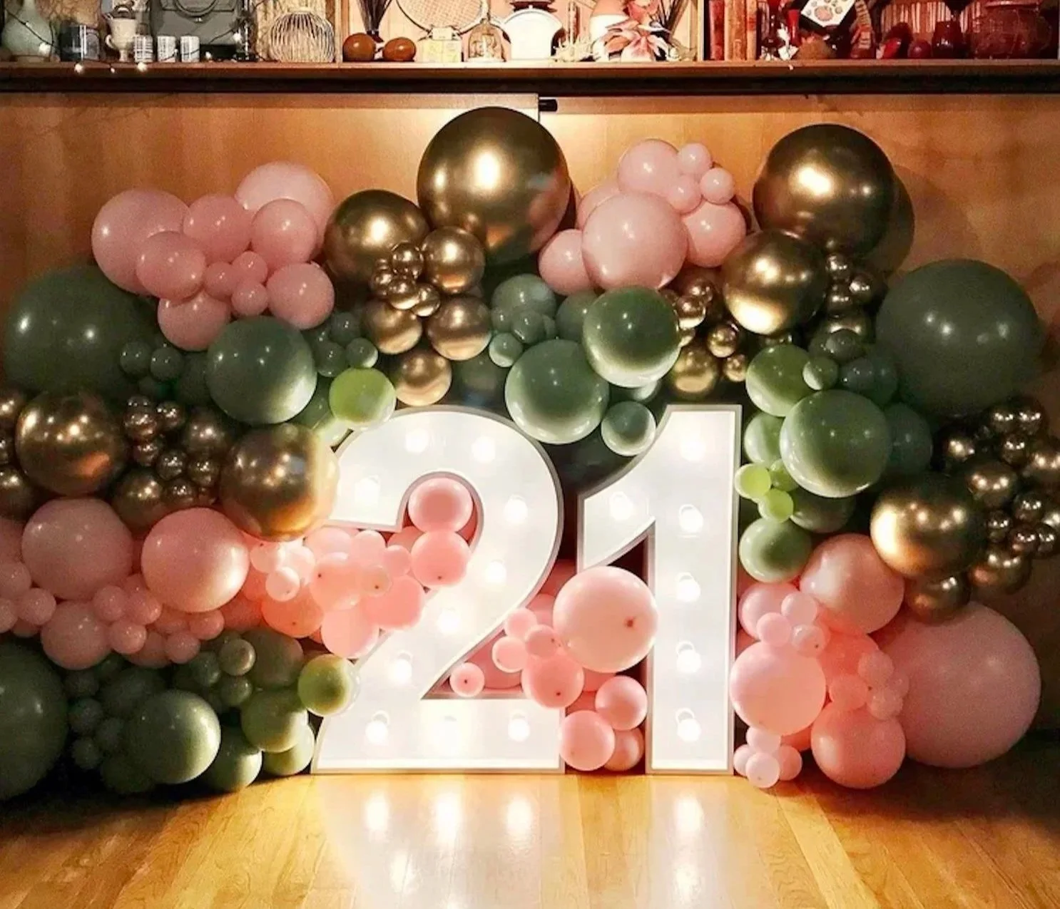 Decorative balloon arrangement with pink, green, and metallic gold balloons surrounding large illuminated white number 21 for a Twenty First birthday party