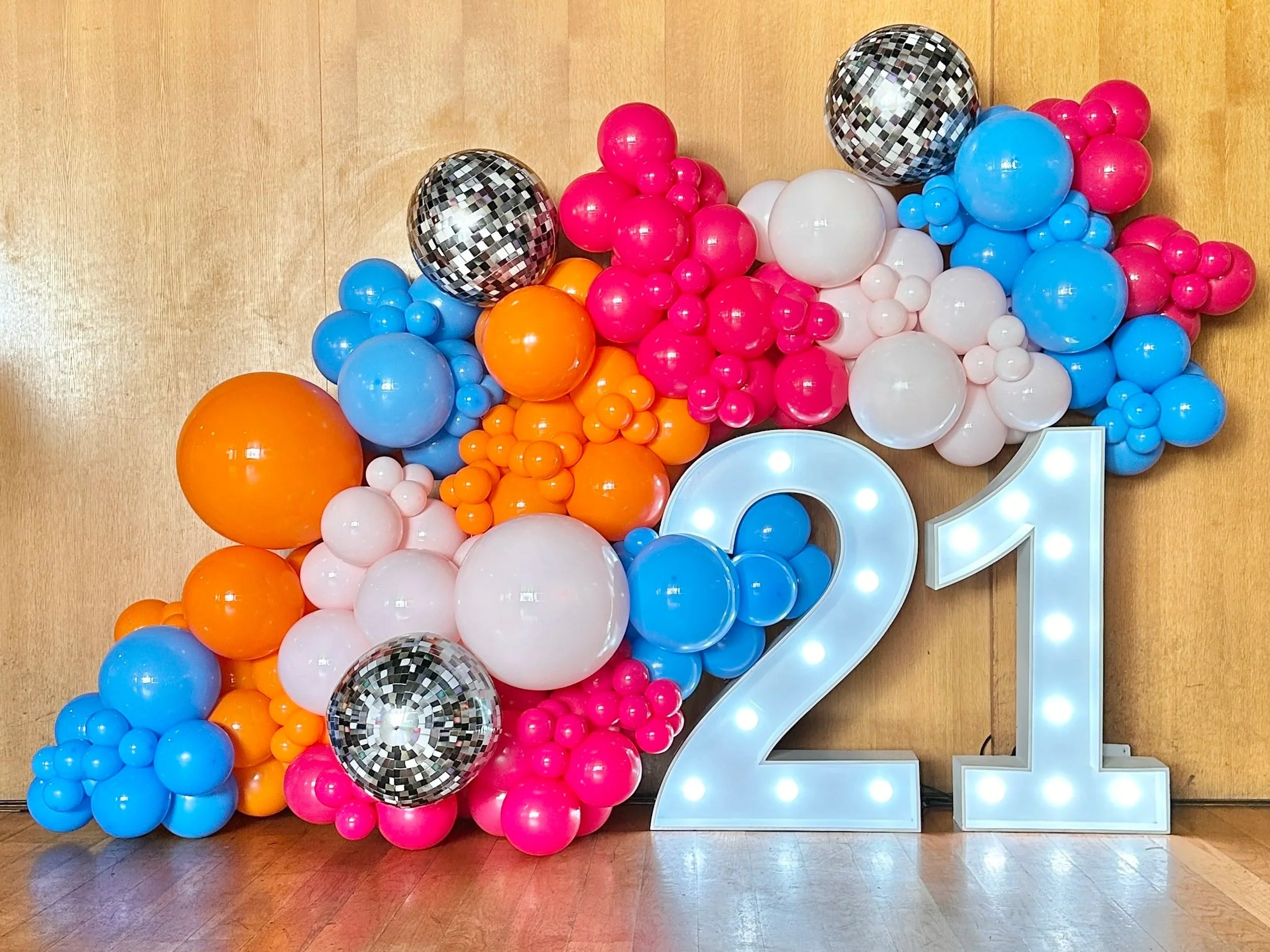 Colourful balloon wall with pink, blue, orange, white, and silver disco ball balloons arranged around a large illuminated number 21