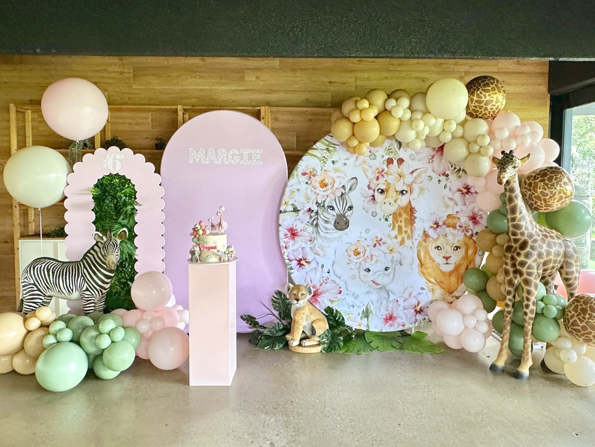 A jungle animal-themed party backdrop with balloons, animal figurines, and animal face illustrations, including a zebra and giraffes.
