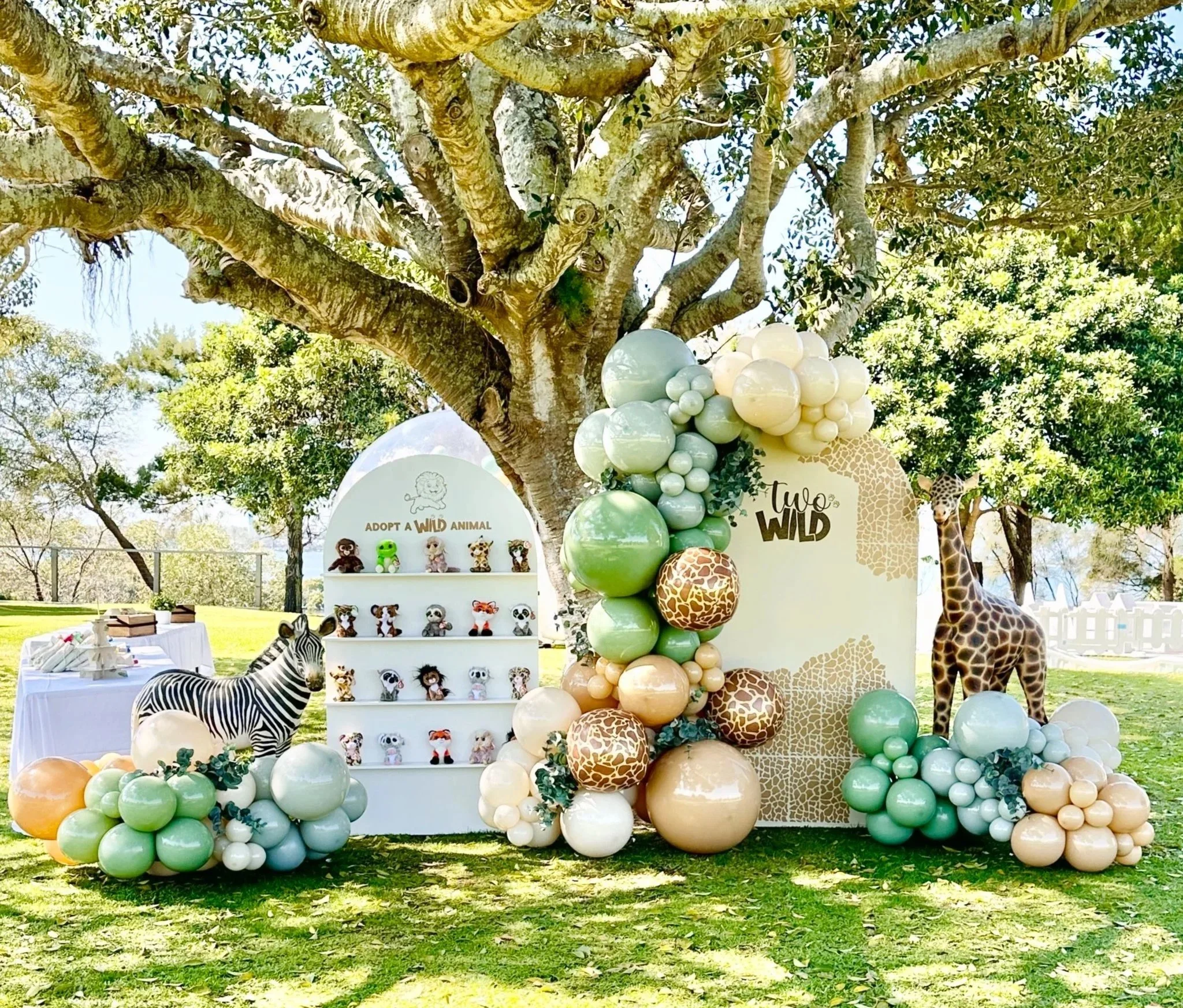 Outdoor jungle-themed event setup with balloons, animal figures, and a decorative backdrop under a large tree.