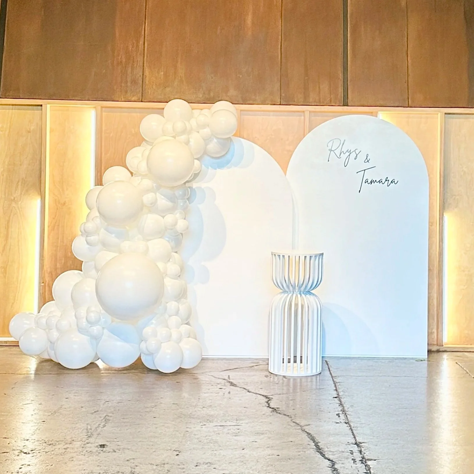 Wedding backdrop with white balloons arranged on the left and a white sign reading 'Rhys & Tamara' on the right, along with a white decorative stand in front, set against a wooden wall.