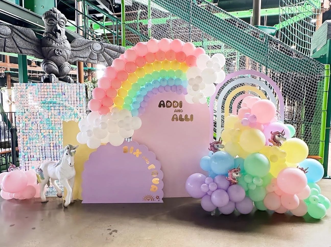 Colorful balloon arrangement with a rainbow and unicorn theme for a celebration at an indoor play area. The backdrop includes pastel-colored blocks with the words "Aodi and Alli" written on it, along with decorative unicorn and rainbow elements.