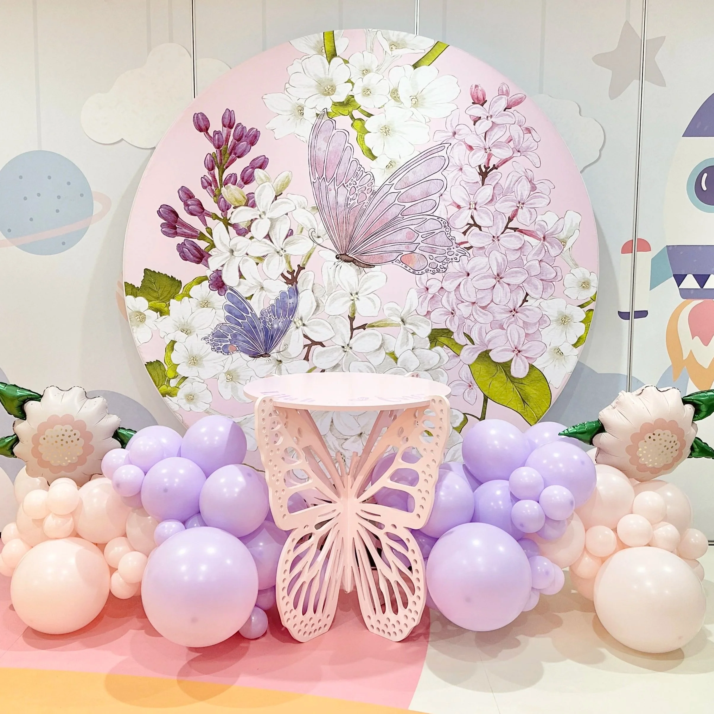 Decorative backdrop with pink, purple, and white flowers, butterflies, surrounded by pink and purple balloons, and a pink butterfly-shaped table in front.
