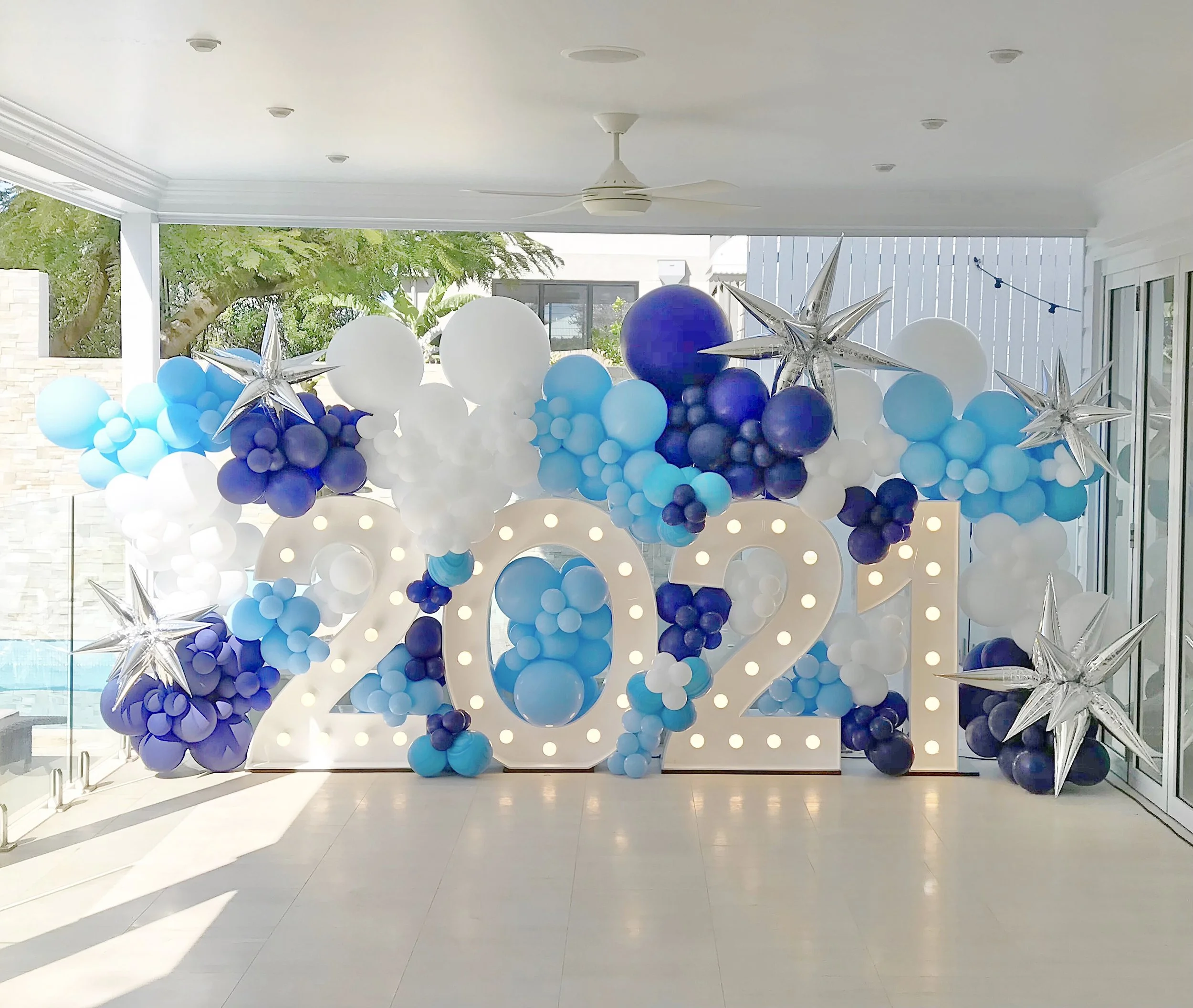 Decorative balloon display with blue, white, and navy balloons, and silver star-shaped balloons, arranged with a large number 23 in the background on a white wall, likely for a celebration or event.