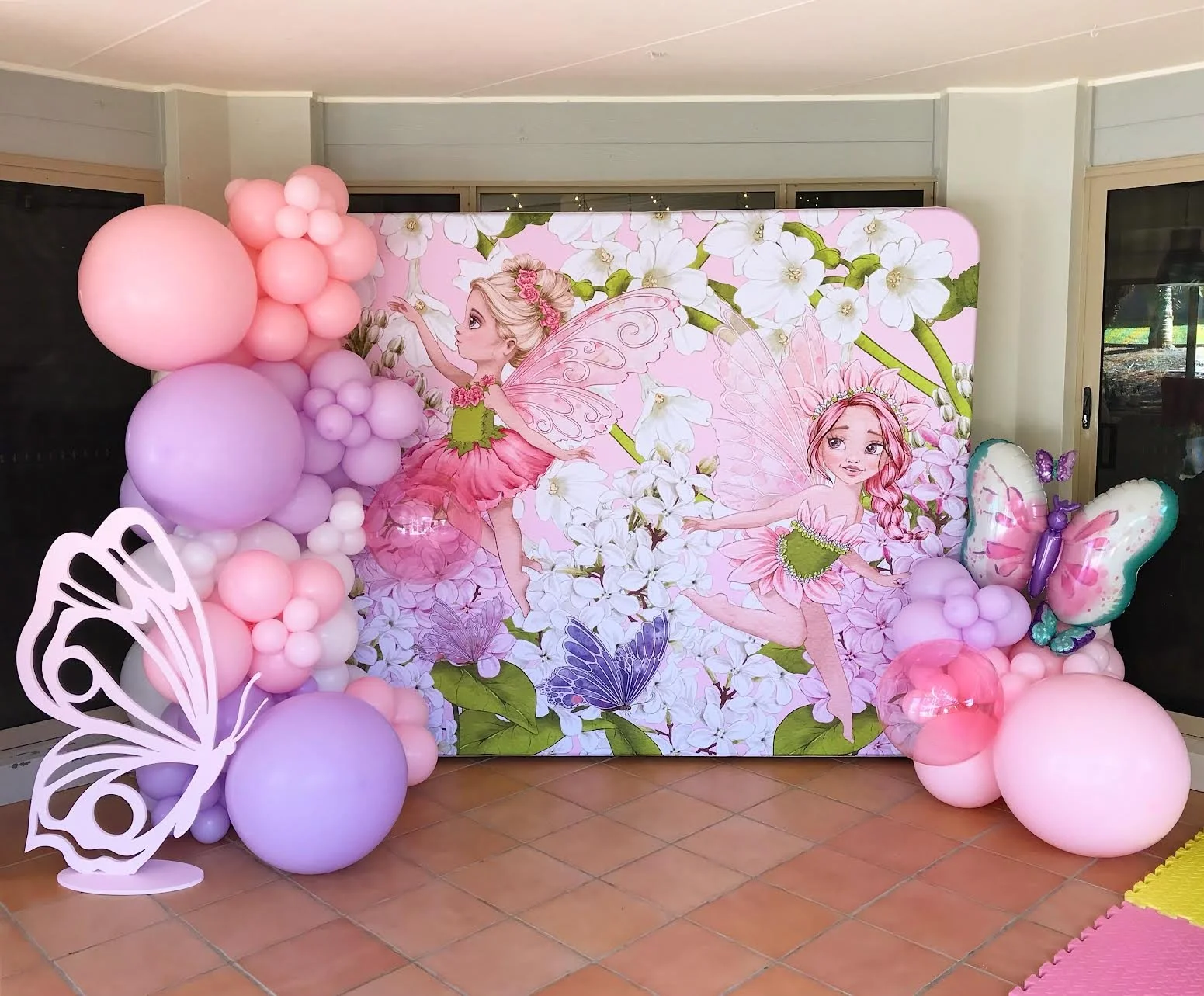 Fairy-themed backdrop with floral and fairy illustrations, pink, purple, and lavender balloons, and decorative butterfly cutouts at a children's party