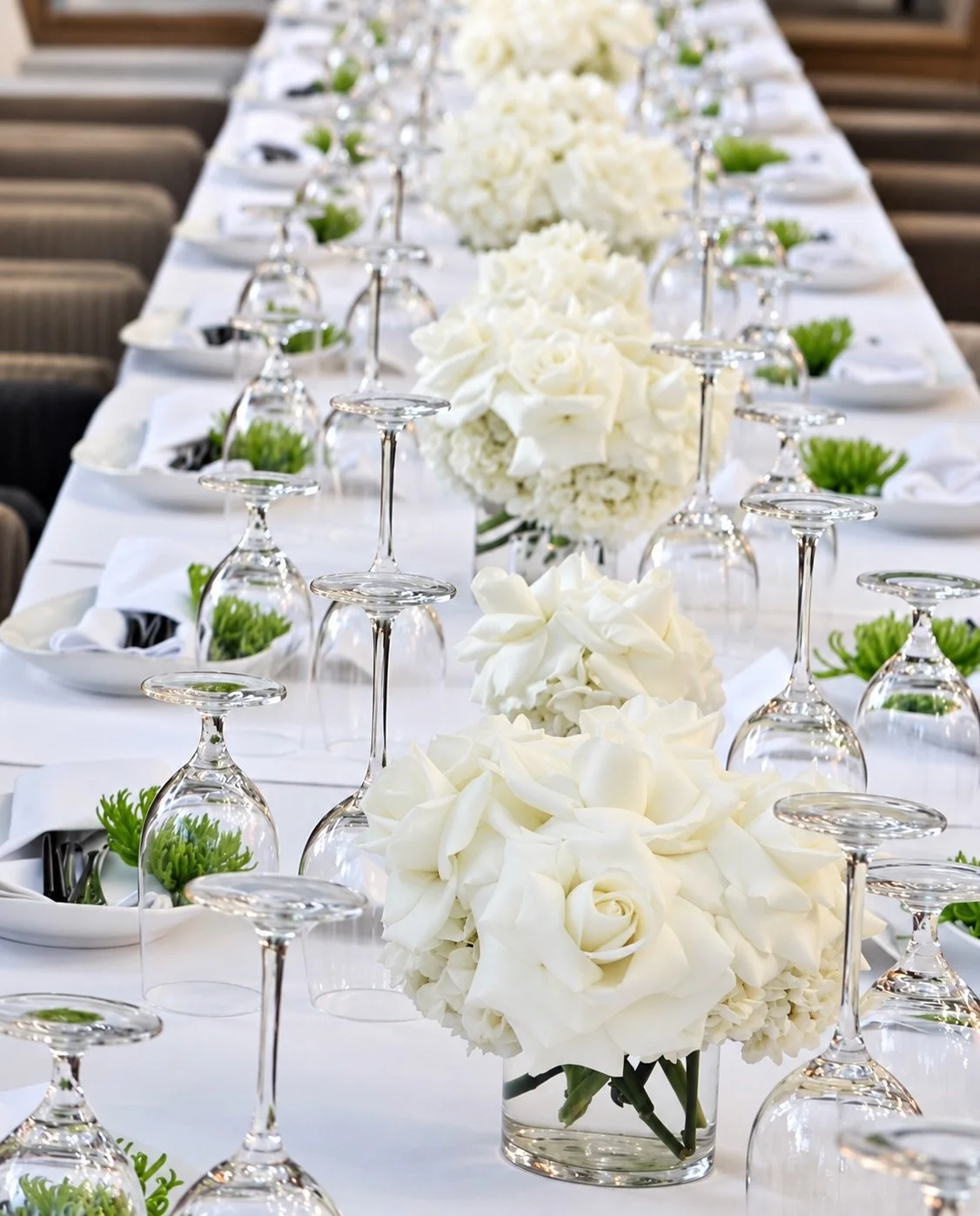 Elegant banquet table with white floral centerpieces, upside-down wine glasses, and small green plants in white bowls, set for a formal event.