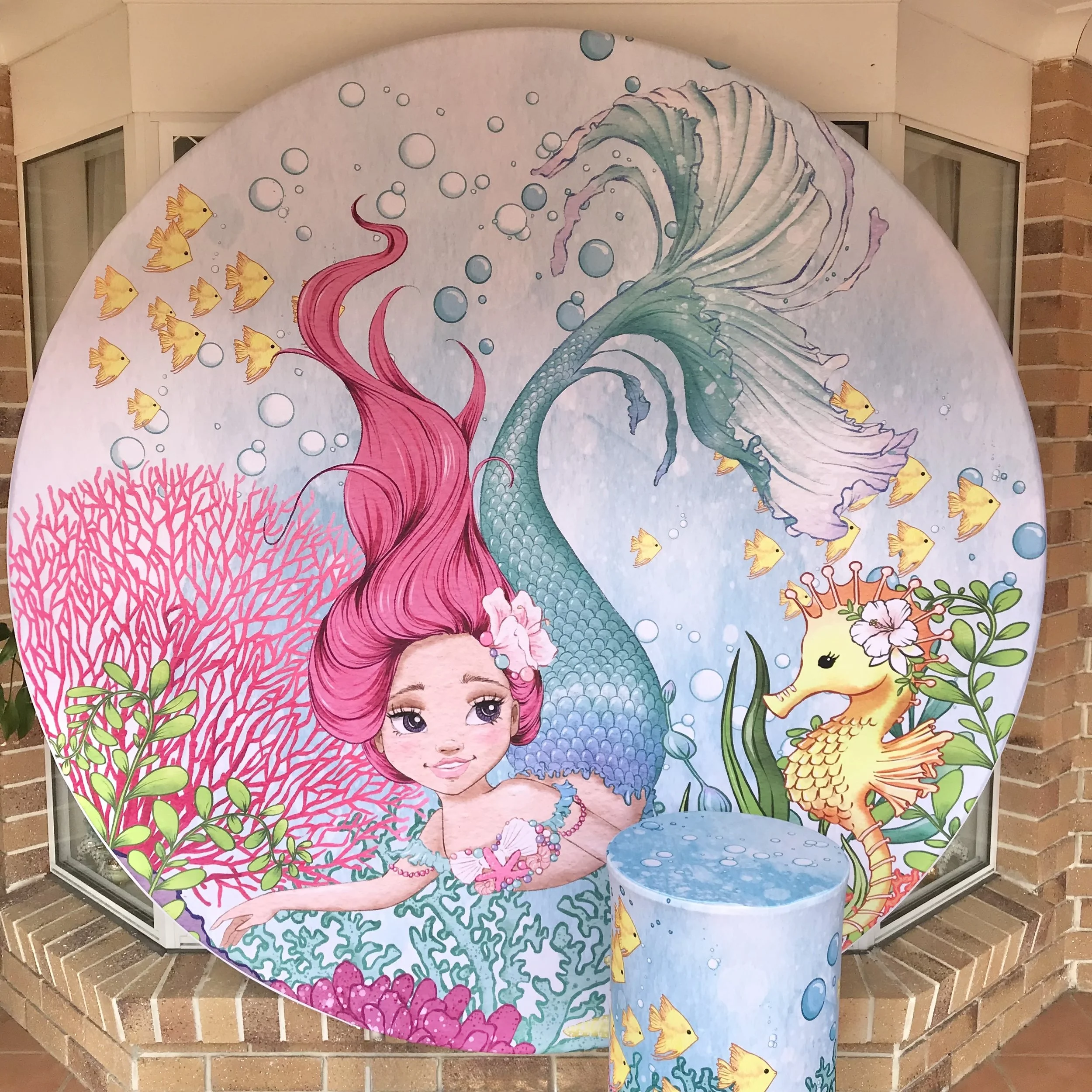 Round mermaid themed backdrop. Mermaid with long pink hair, underwater and surrounded by sea creatures like seahorses, fish, and coral