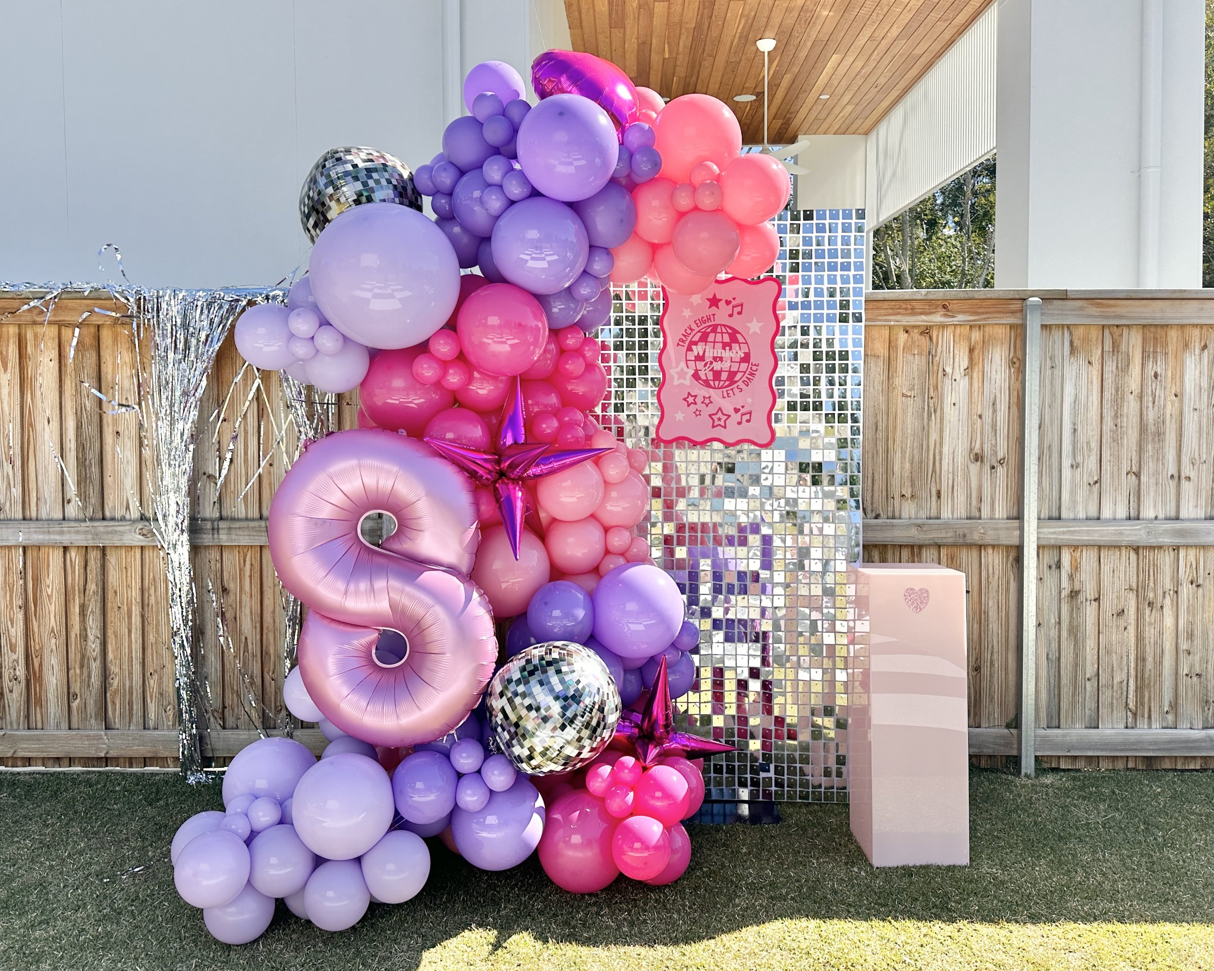 Decorative balloon display with pink and purple balloons, a disco ball, silver tinsel, a pink sign with music and dance theme, set against a driveway and wooden fence with a wooden roof ceiling.