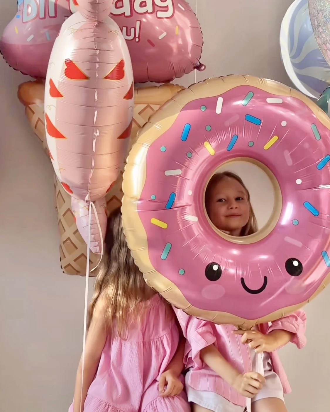 Child holding a large pink donut-shaped balloon with a smiling face, surrounded by other balloons including a donut and a character ice cream cone, at a birthday celebration.