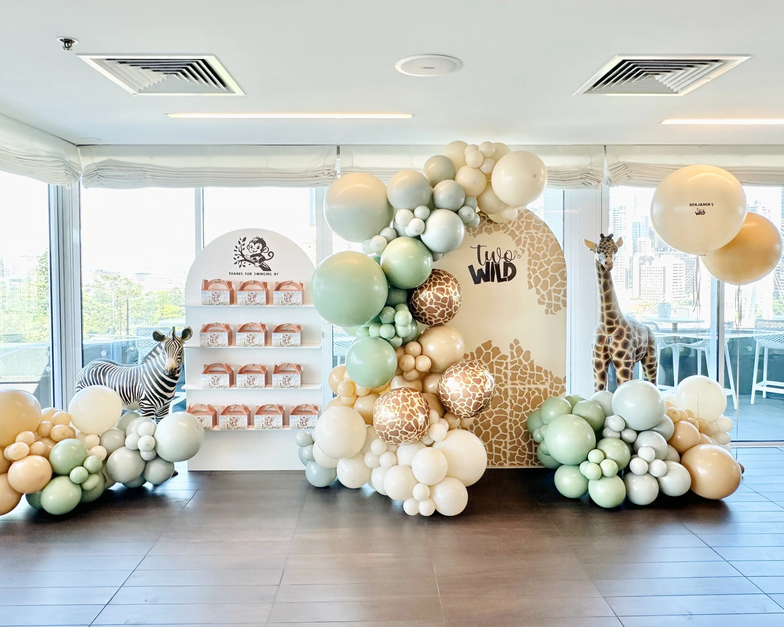 Zoo-themed baby shower display with zebra and giraffe balloons, a balloon arch, and a giraffe cutout, featuring decorations with animal print patterns and greenery.