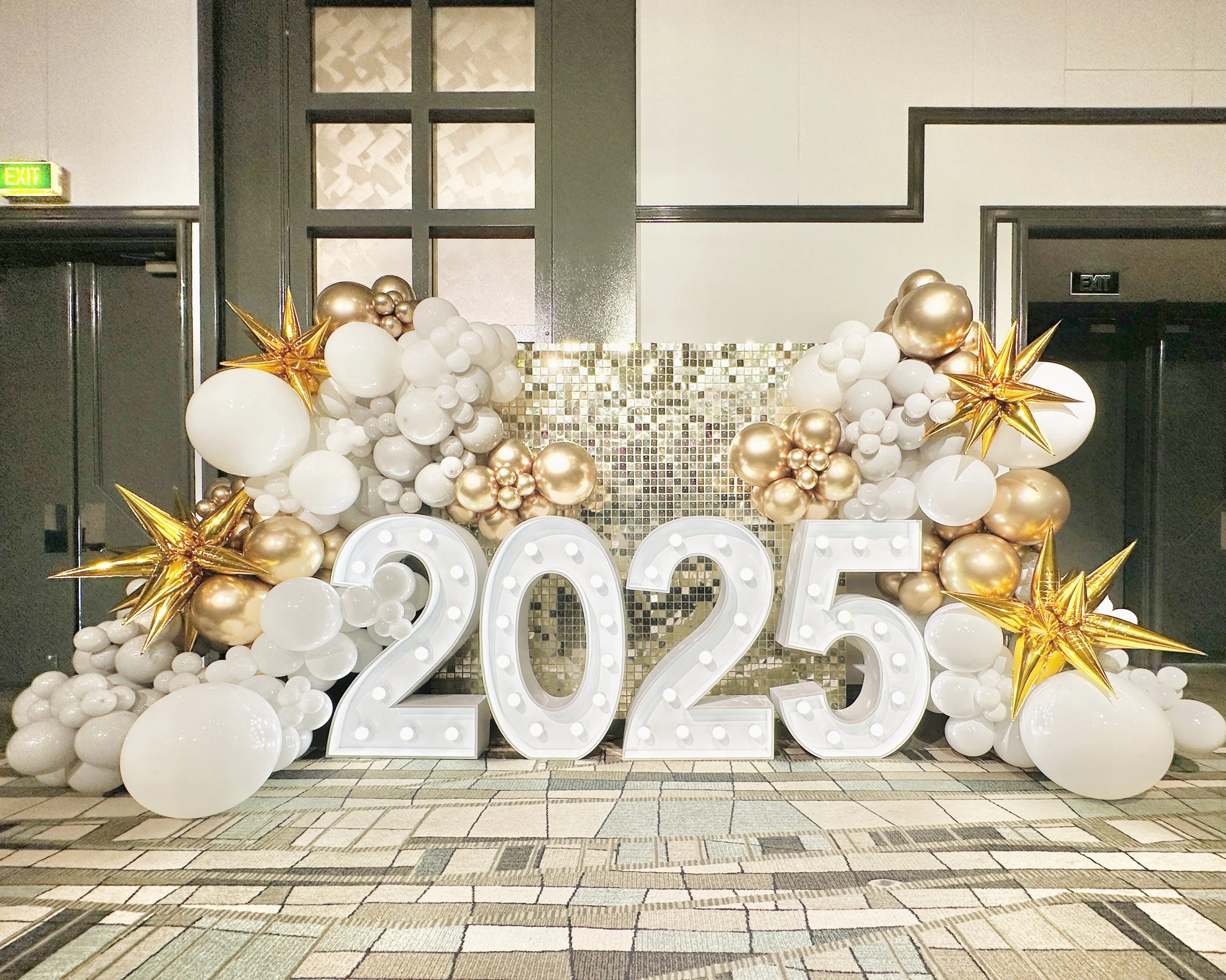 2025 illuminated numbers,  surrounded by white and gold balloons set against a gold shimmer wall backdrop