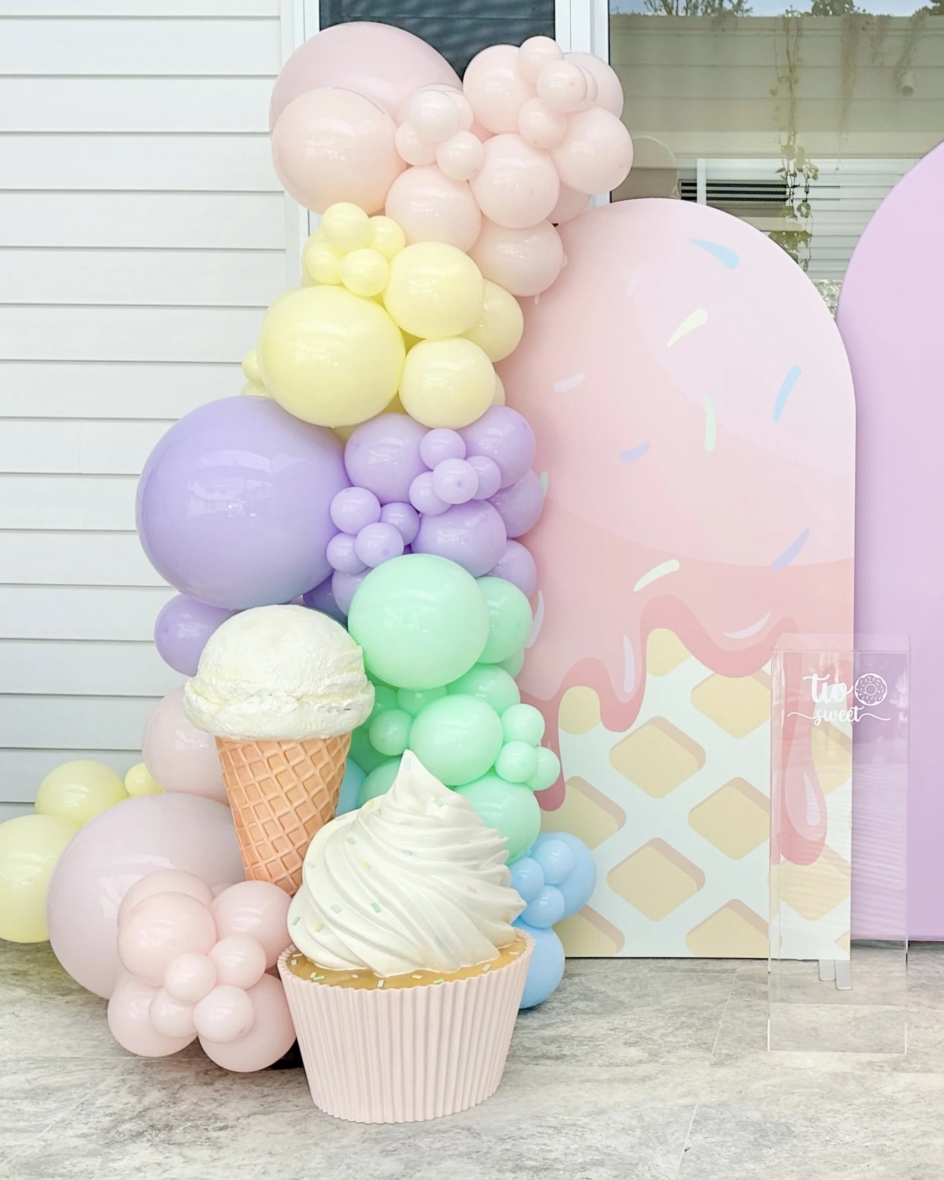 Decorative pastel-colored balloon arrangement and cupcake prop and ice cream prop, set against a pink backdrop with sprinkles and a clear acrylic cake sign that says 'two sweet'.