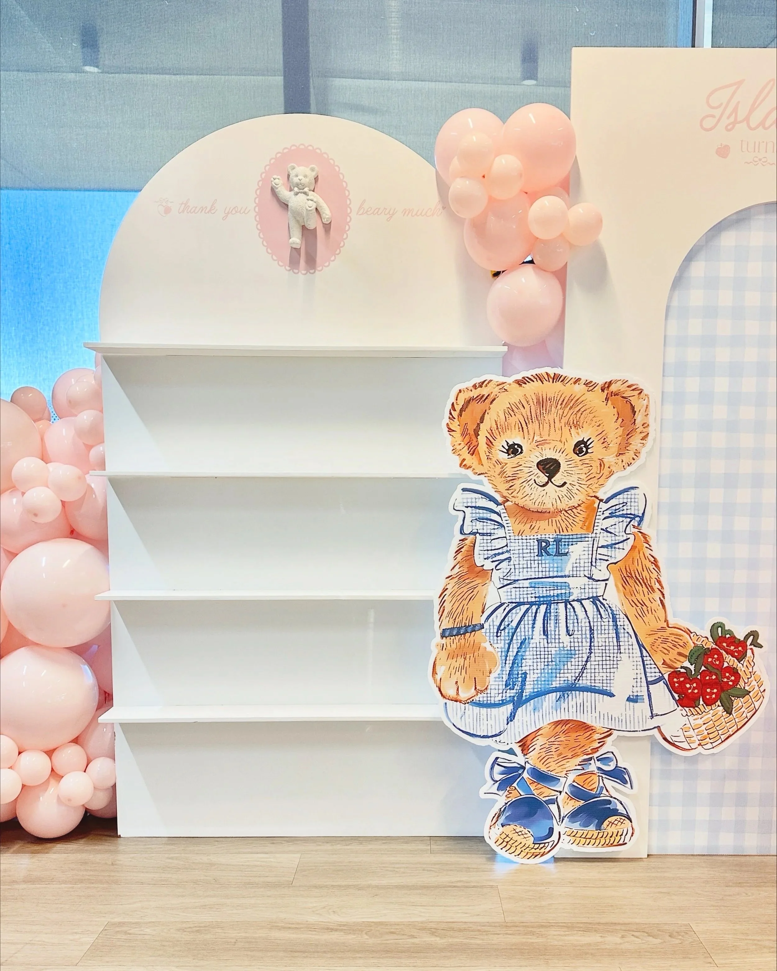 A decorative display with pink balloons on the left, a white shelf in the middle, and a large cutout of a cute teddy bear wearing a striped apron holding a basket of strawberries on the right. The background features a blue and white checkered pattern, and the floor is wood.