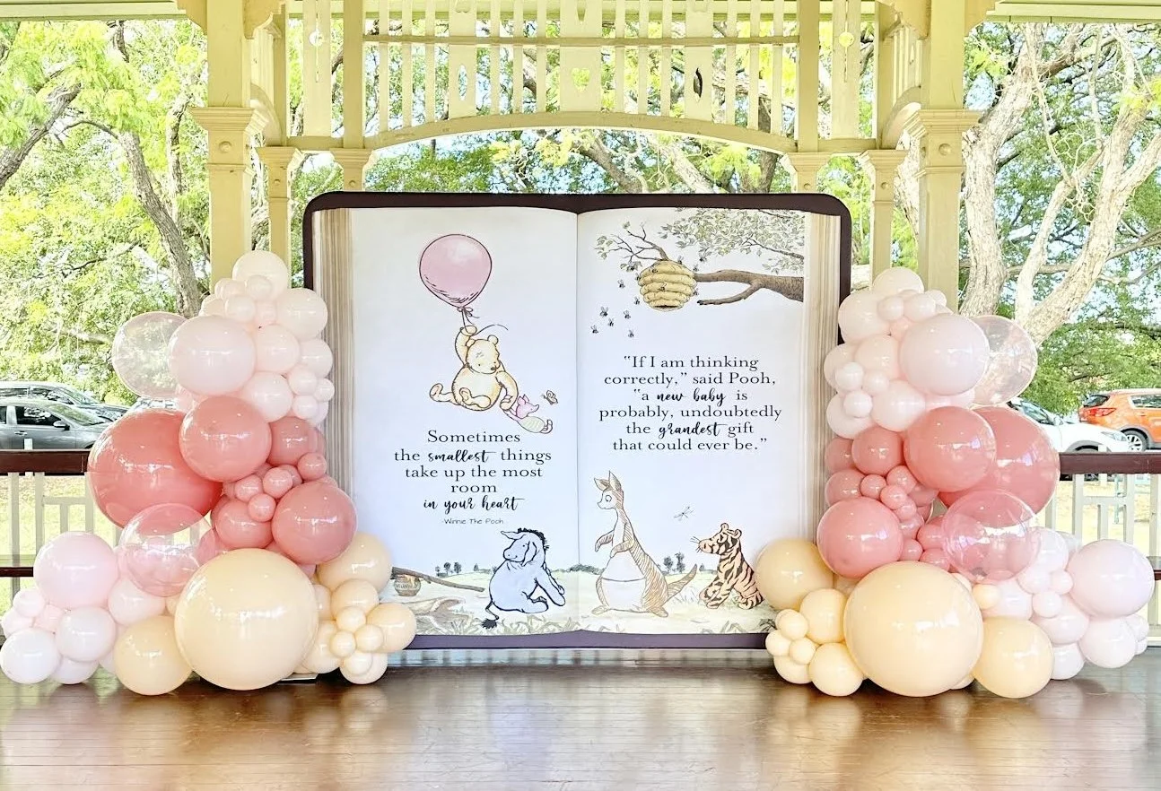 Decorative display with a large open storybook featuring Winnie the Pooh characters, surrounded by pink, white, and cream-colored balloons, set outdoors with trees and cars in the background.