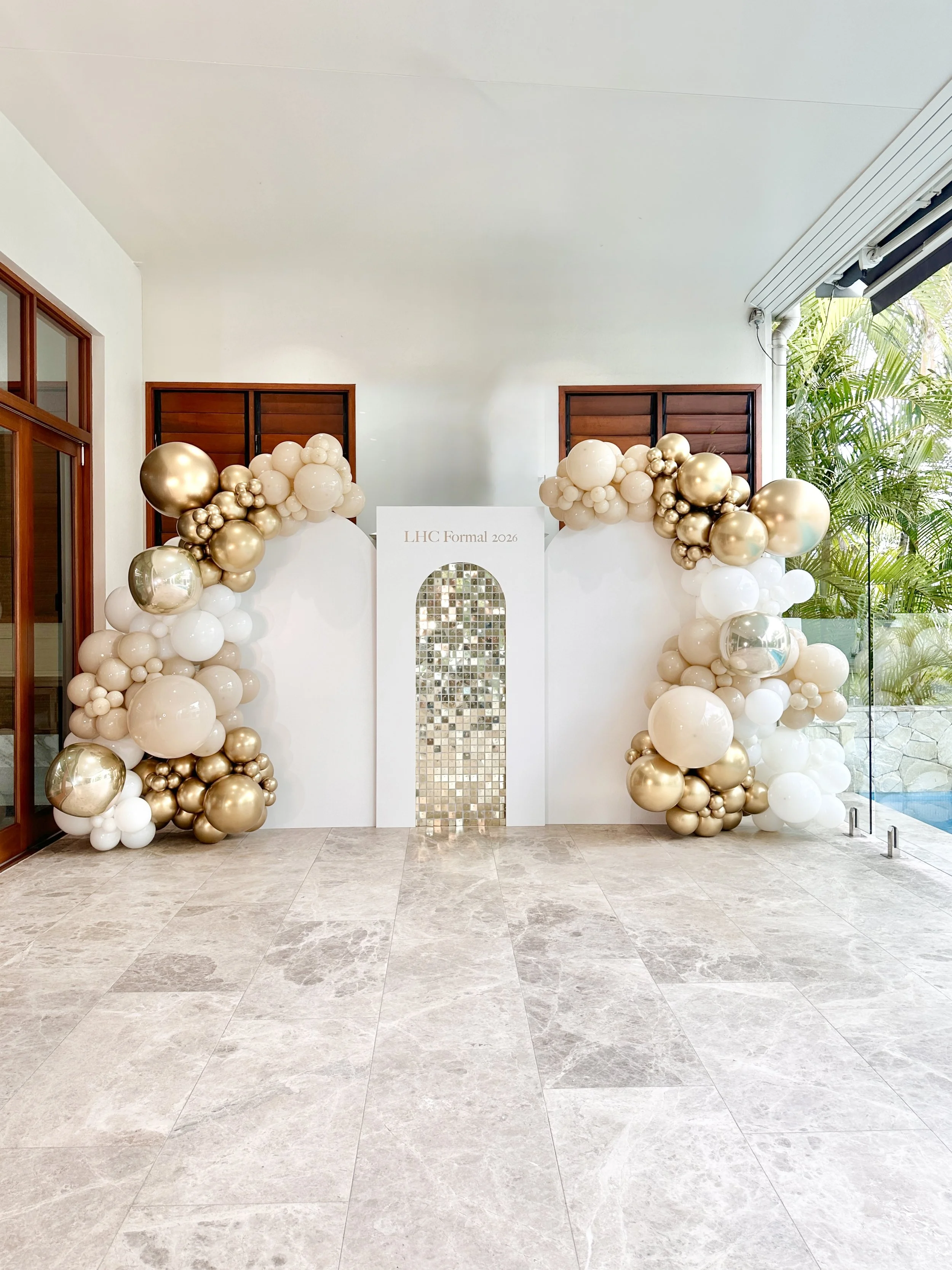 Decorative balloon arrangement with white, gold, and champagne-colored balloons, and a white backdrop featuring a sign that reads 'LHC Formal 2026,' set in a bright indoor area with large tiles and greenery outside.