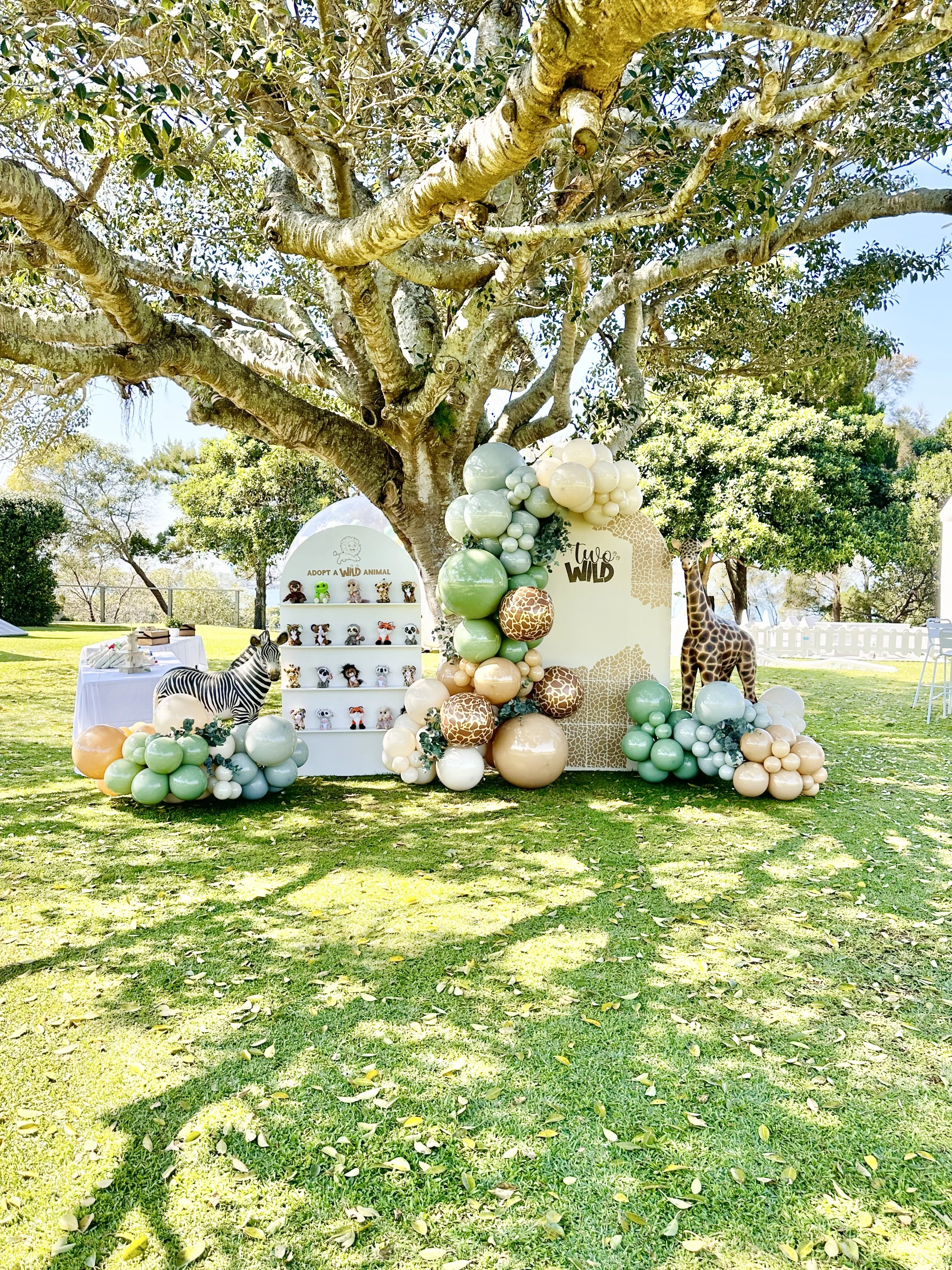 Outdoor jungle-themed event setup with balloons, animal figures, and a decorative backdrop under a large tree.