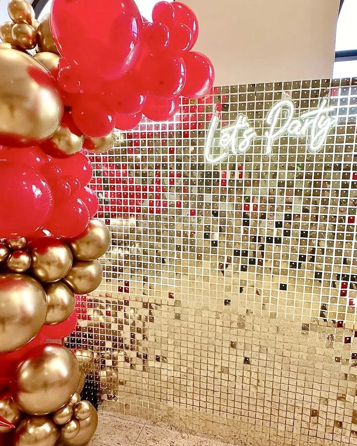 Gold shimmer backdrop, red and gold balloons and a neon sign which reads Let's Party