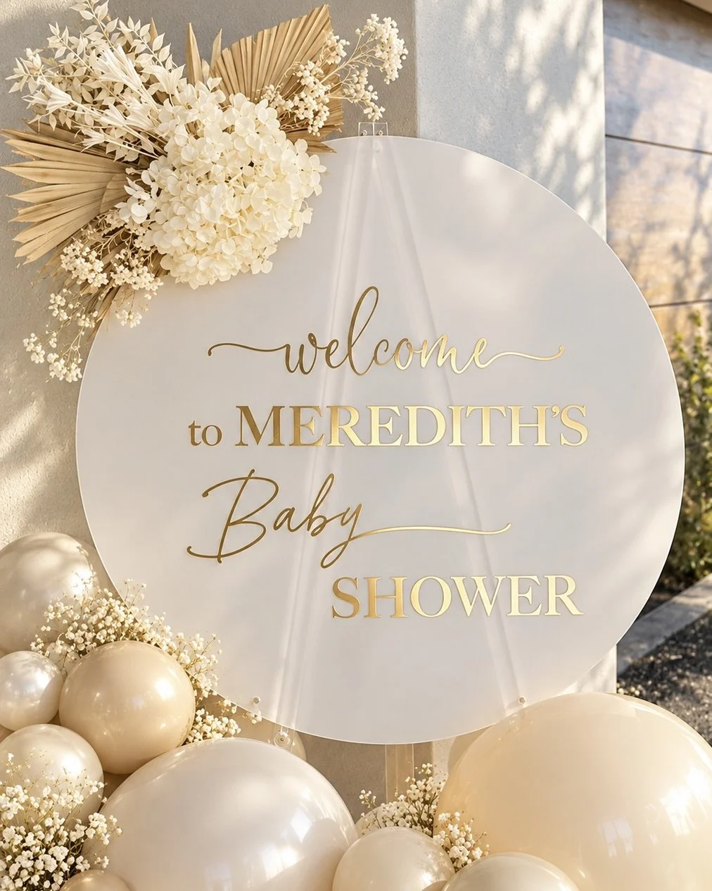 Decorative baby shower sign framed by green, white, and beige balloons with eucalyptus leaves. The sign reads 'Celebrating Baby Smart, a little one is on the way,' near large glass doors showing outdoor greenery.
