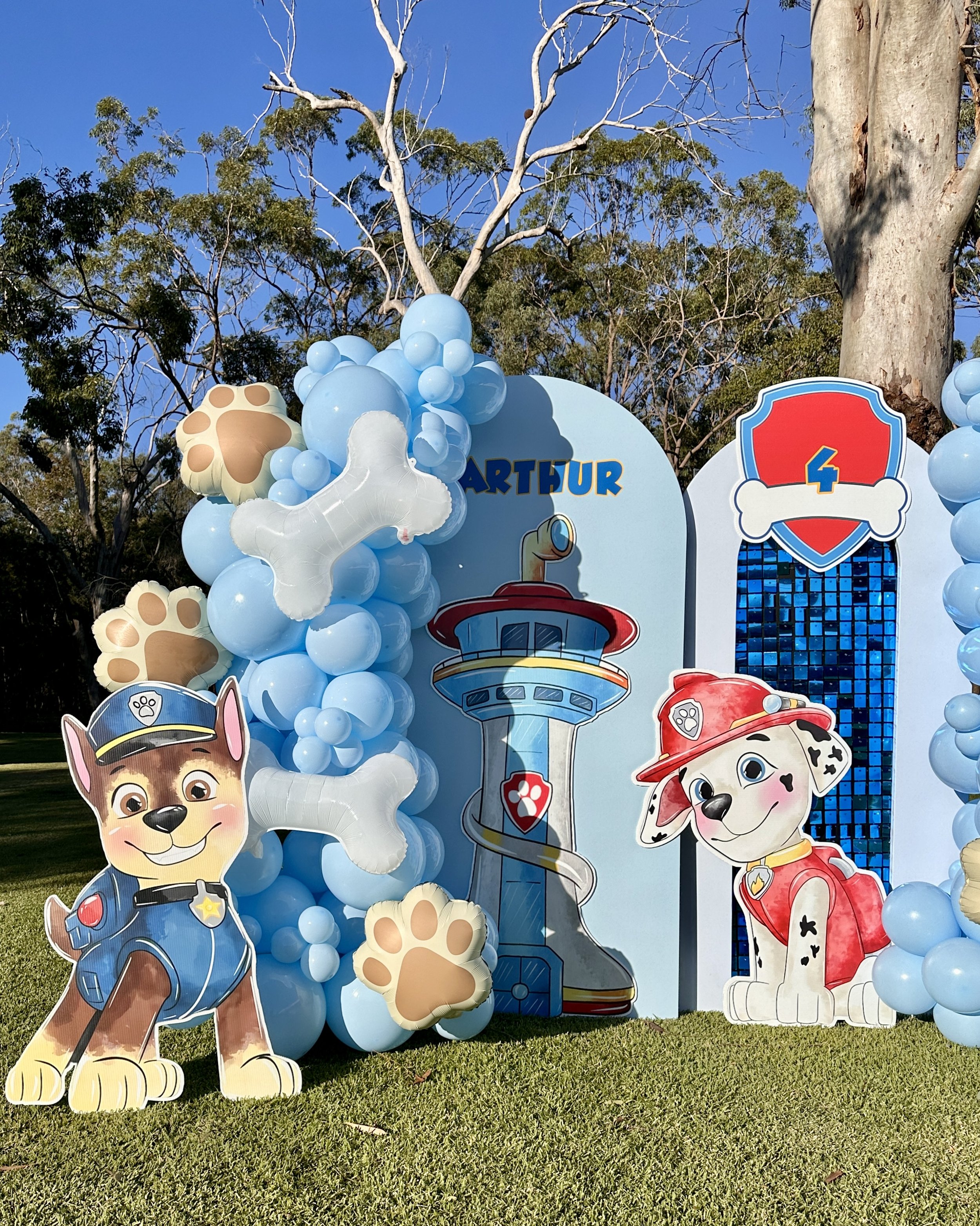 Decorations featuring characters from Paw Patrol, including Chase and Marshall, with balloons and a sign reading "Arthur" and a badge with the number 4, set outdoors with trees and grass in the background.