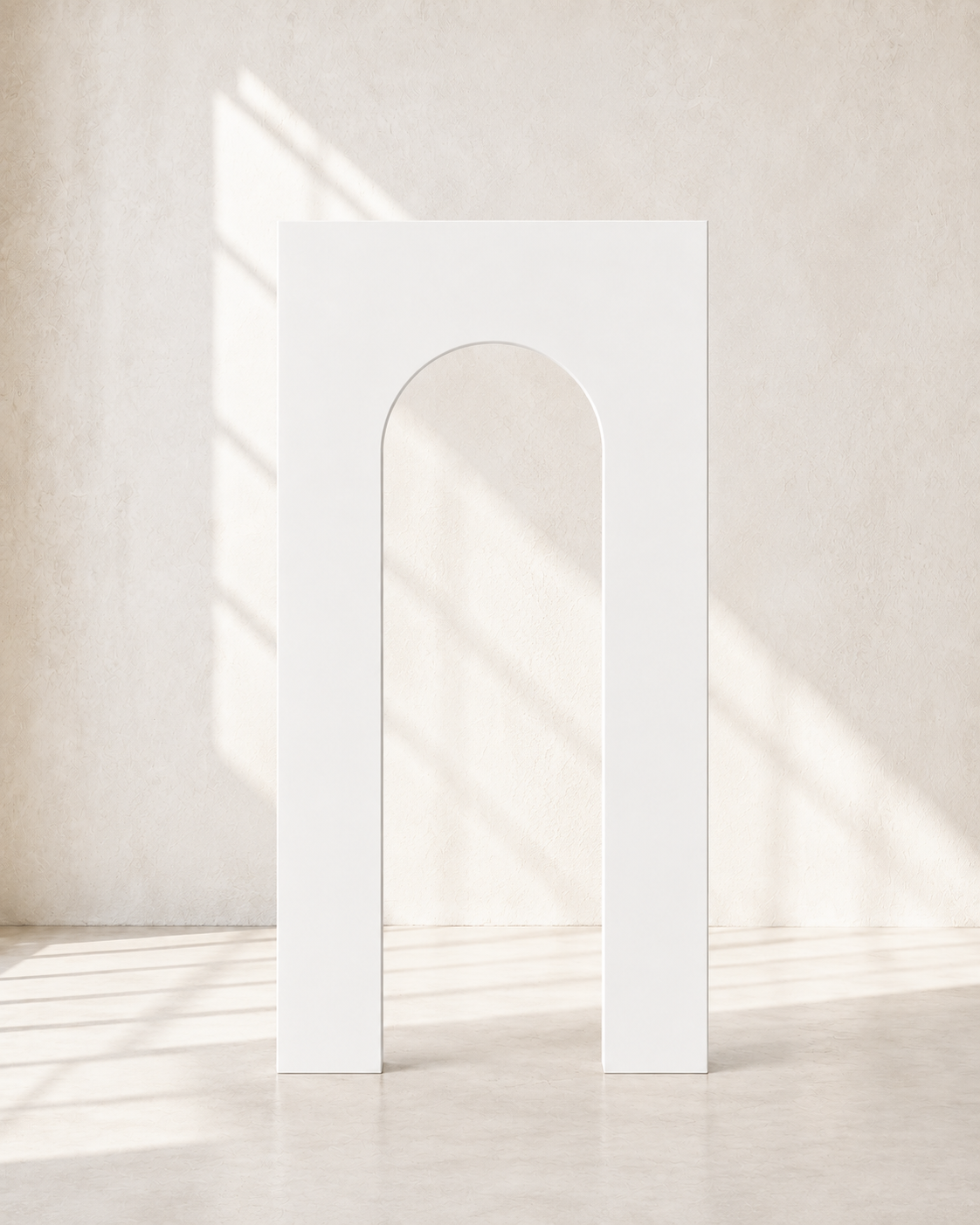 Minimalist white arch-shaped sculpture or doorway standing in a bright, neutral room with sunlight and shadows on the wall.