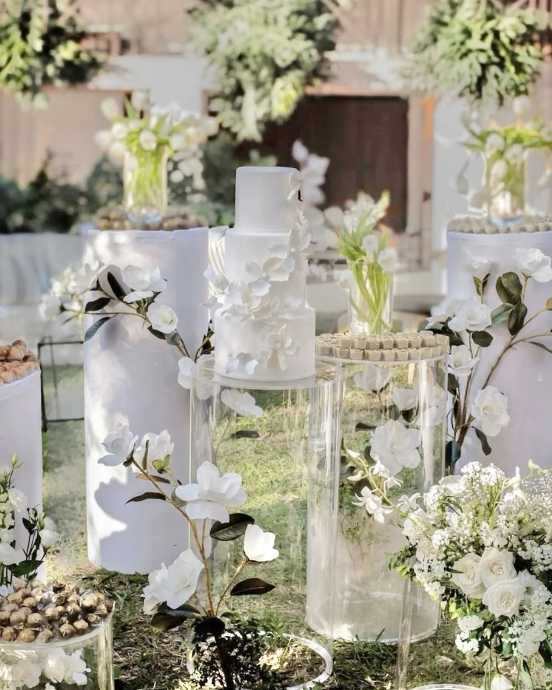 Elegant outdoor table decorated with white floral arrangements, vases, and a multi-tiered white cake, suggestive of a wedding or celebration event.