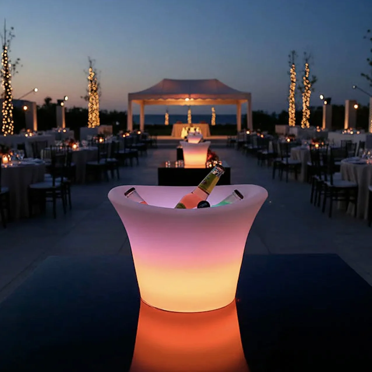 Outdoor reception area at dusk with illuminated ice bucket featuring bottles of beer or wine in the foreground, and tables with candles and chairs arranged in the background under string lights and a canopy, overlooking a body of water.