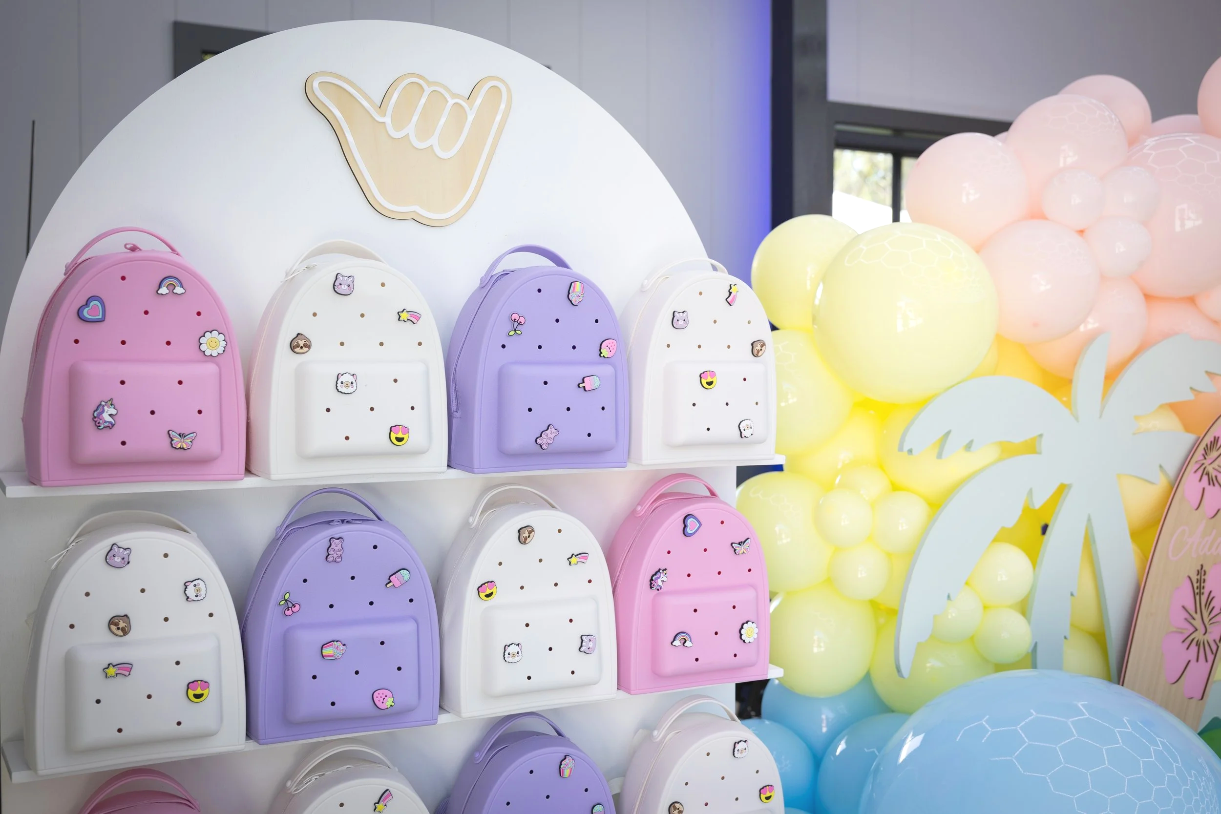 Colorful display of small pastel-colored backpacks with cute sticker decorations, positioned in front of a backdrop of yellow, pink, and blue balloons.