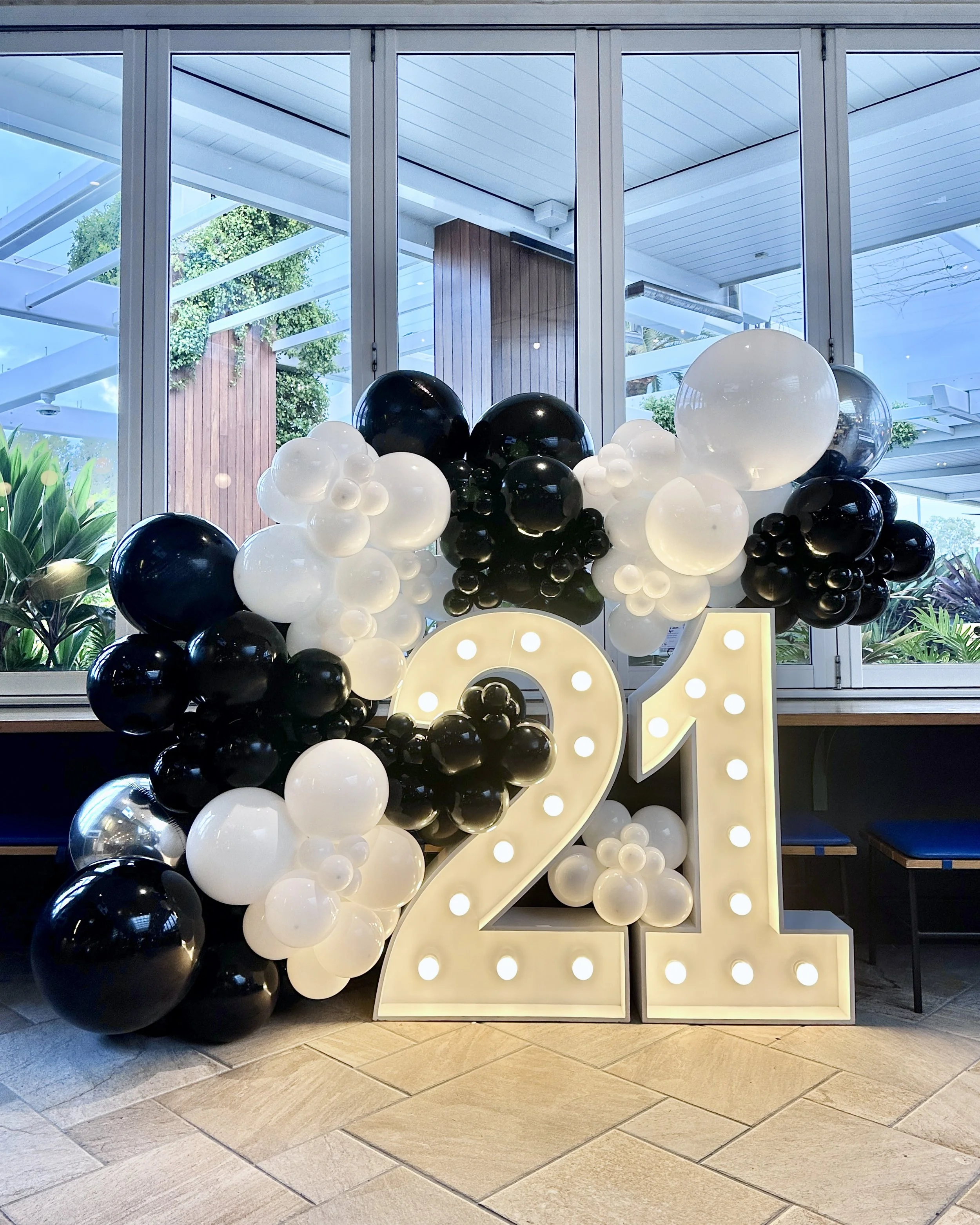 Decorative display of black and white balloons arranged around illuminated letter signs spelling '2024' against a large window with a view of trees outside.