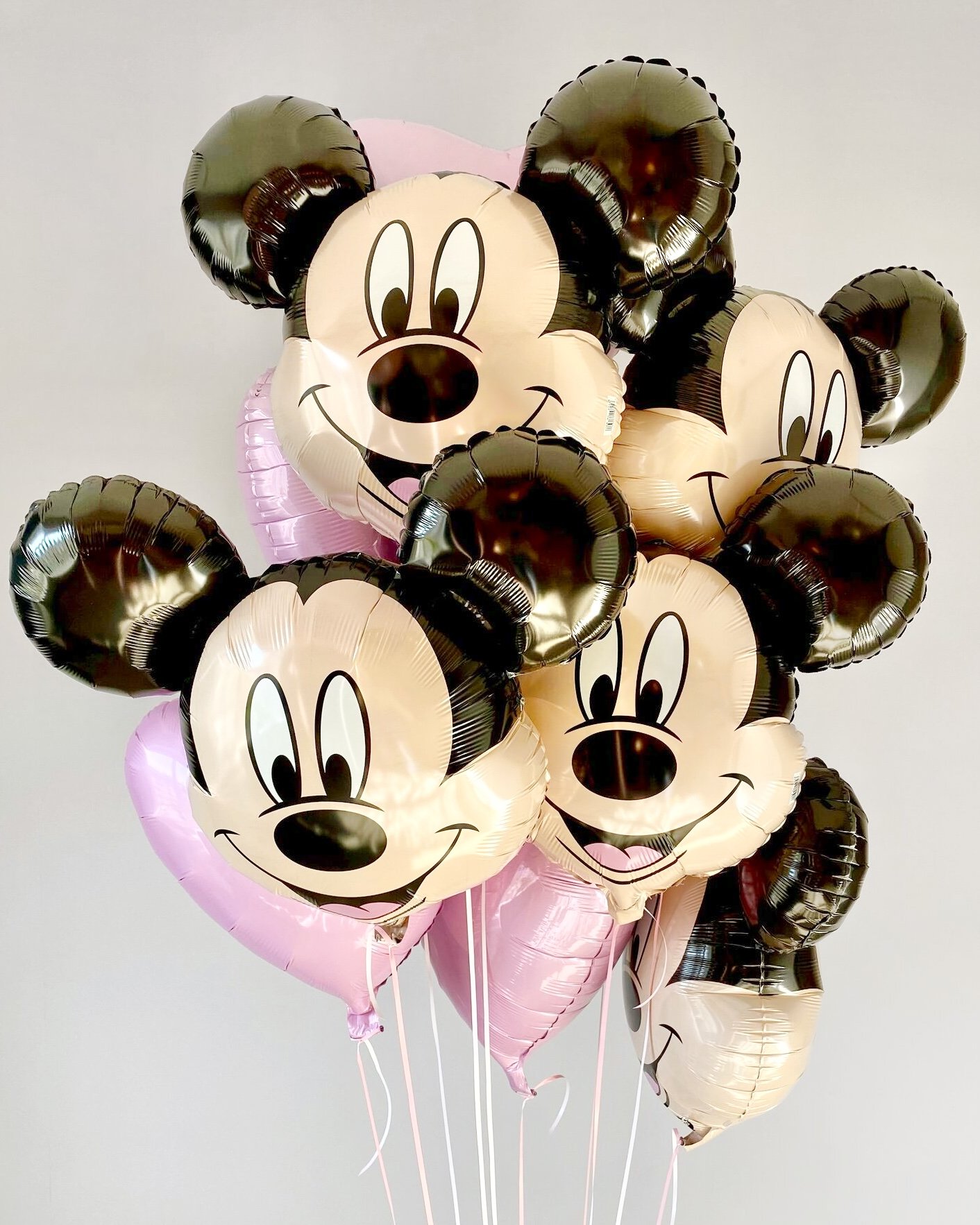 Cluster of Mickey Mouse and Minnie Mouse themed balloons with smiling faces, black ears, and pink and beige colors, tied together against a plain background.